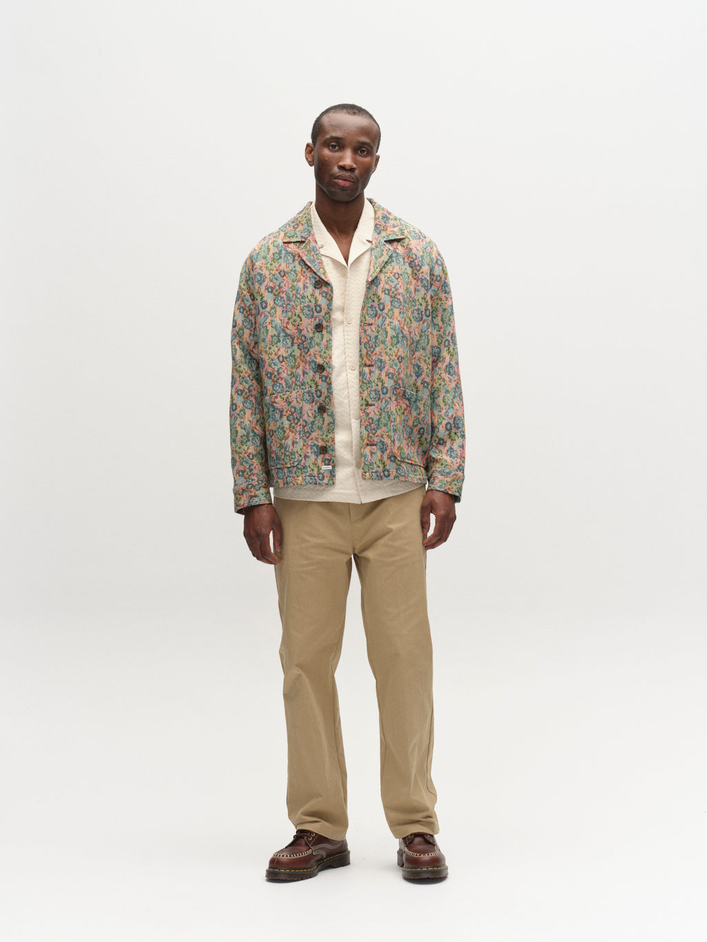 A man stands against a white background wearing the Gabba GABaustin POKT Kscope LS Overshirt - Abstract Flowers, layered over a cream shirt with beige pants and brown shoes, embodying effortless seasonal style.