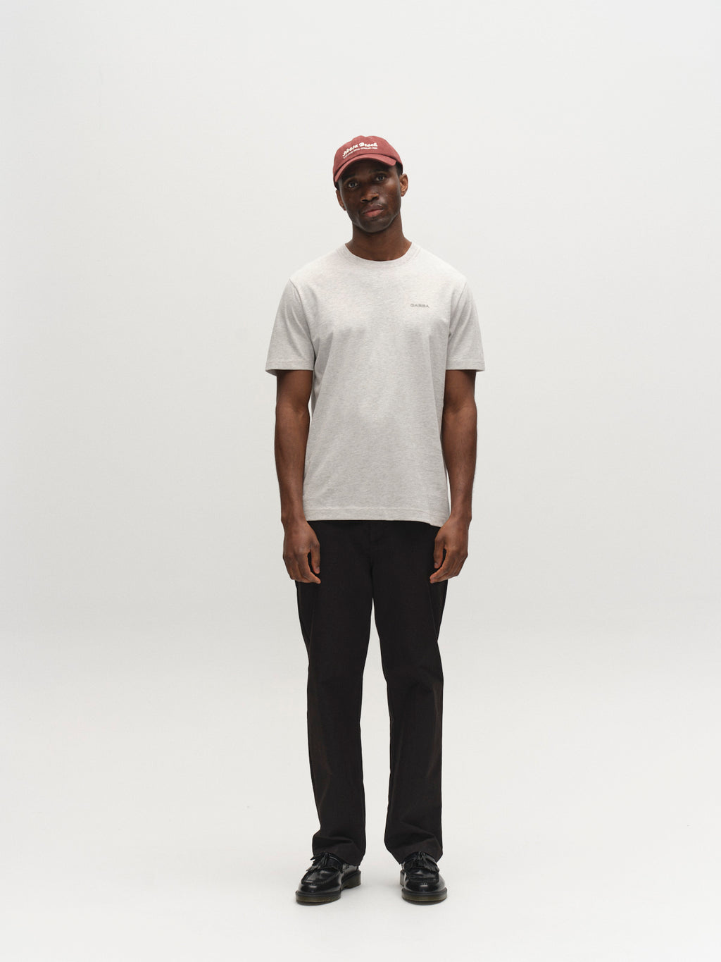 A man stands against a plain white background, wearing the Gabba GABdune Melange New SS T-Shirt in Lt. Grey Mel, black pants and shoes, and a red cap. He faces forward with a neutral expression and relaxed posture.