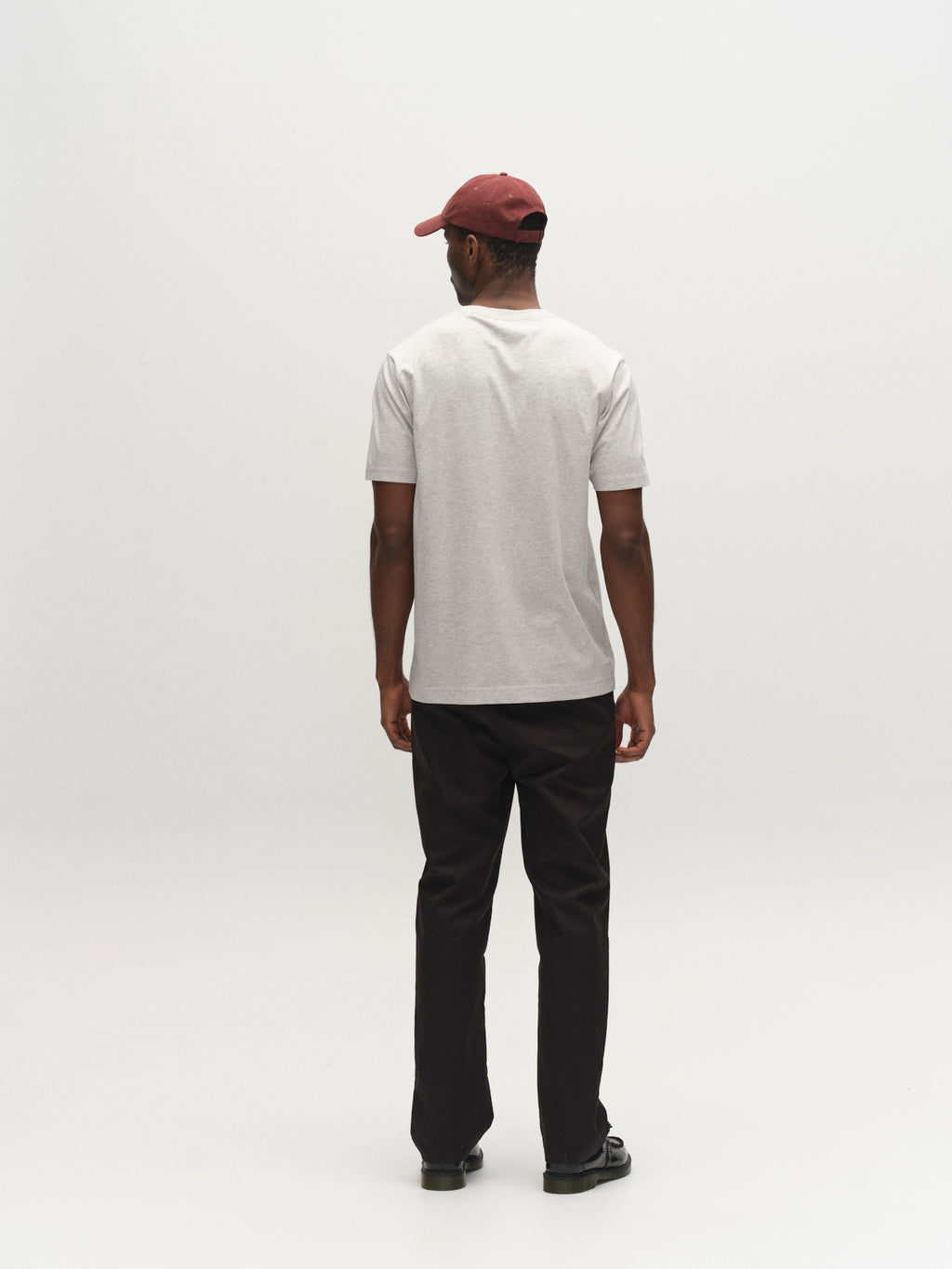 A person in a Gabba GABdune Melange New SS T-Shirt - Lt. Grey Mel faces away from the camera, paired with dark pants, black shoes, and a maroon cap, set against a plain light background.