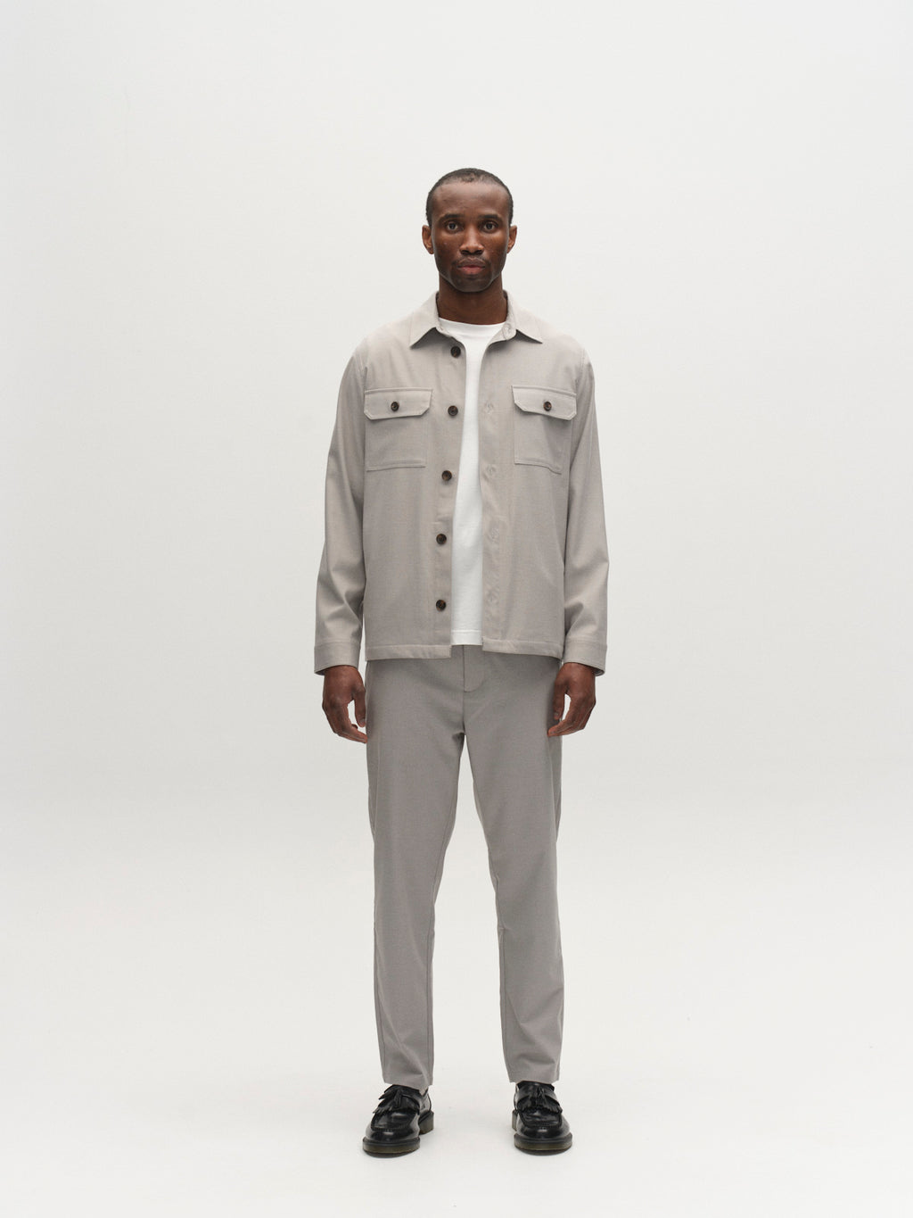 A man stands in a minimalist studio wearing the Gabba GABmonza Tasca Deme Pant in Mineral Gray with an elastic waistband, paired with a light gray button-up jacket, a white shirt, and black shoes against a plain white background.