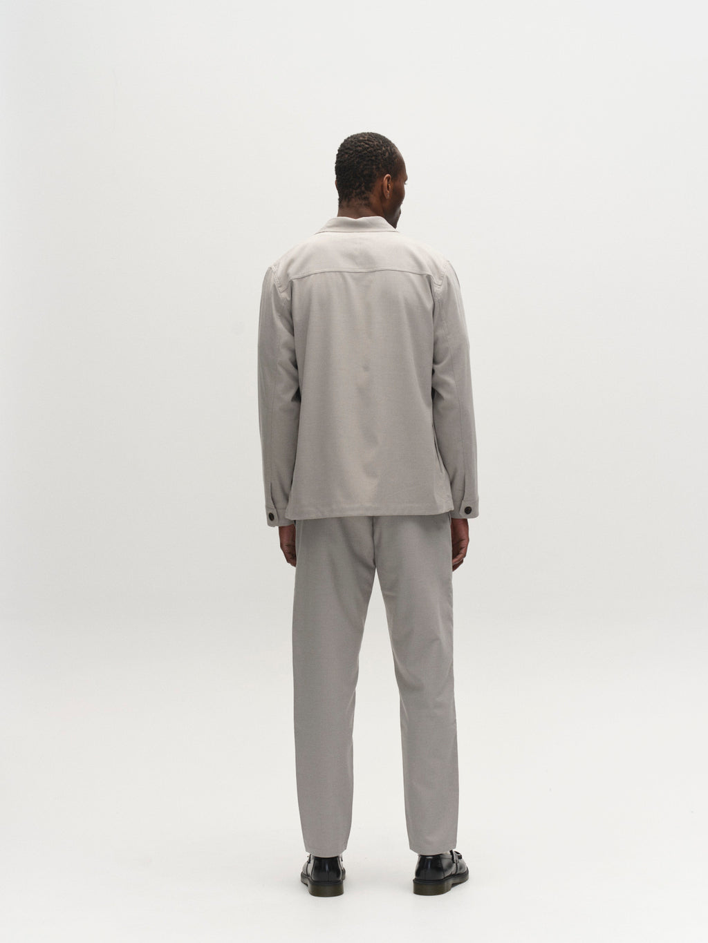 A man faces away from the camera against a plain white background, wearing Gabba's GABmonza Tasca Deme Pant in Mineral Gray with an elastic waistband, a matching suit jacket, and black shoes.