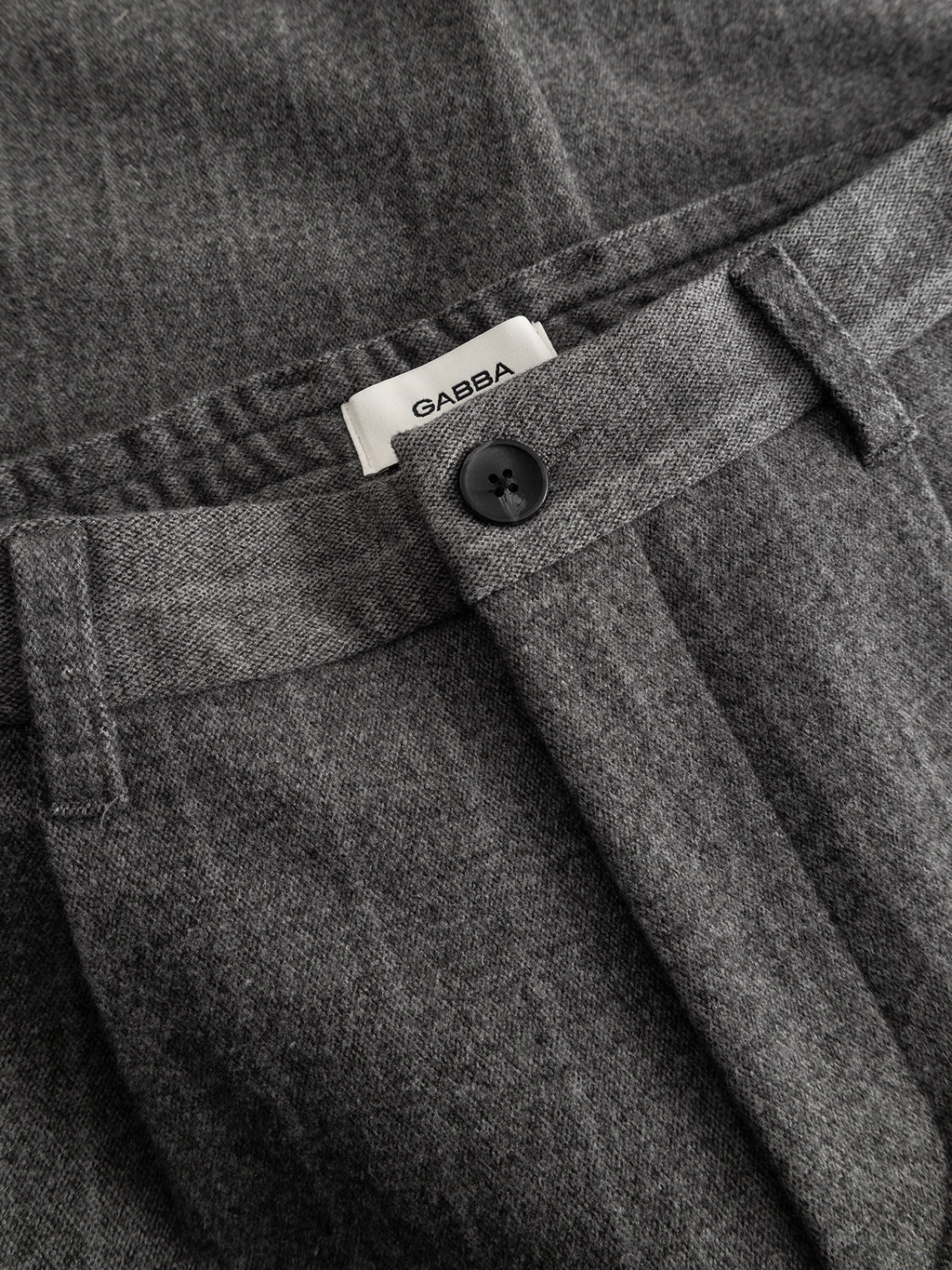 Close-up of Gabba Napoli 19 AX Stripes pants in dark grey, showcasing a relaxed fit with a black button, belt loops, and chino-style pockets. The inside waistband features a white label with the Gabba brand name printed on it.