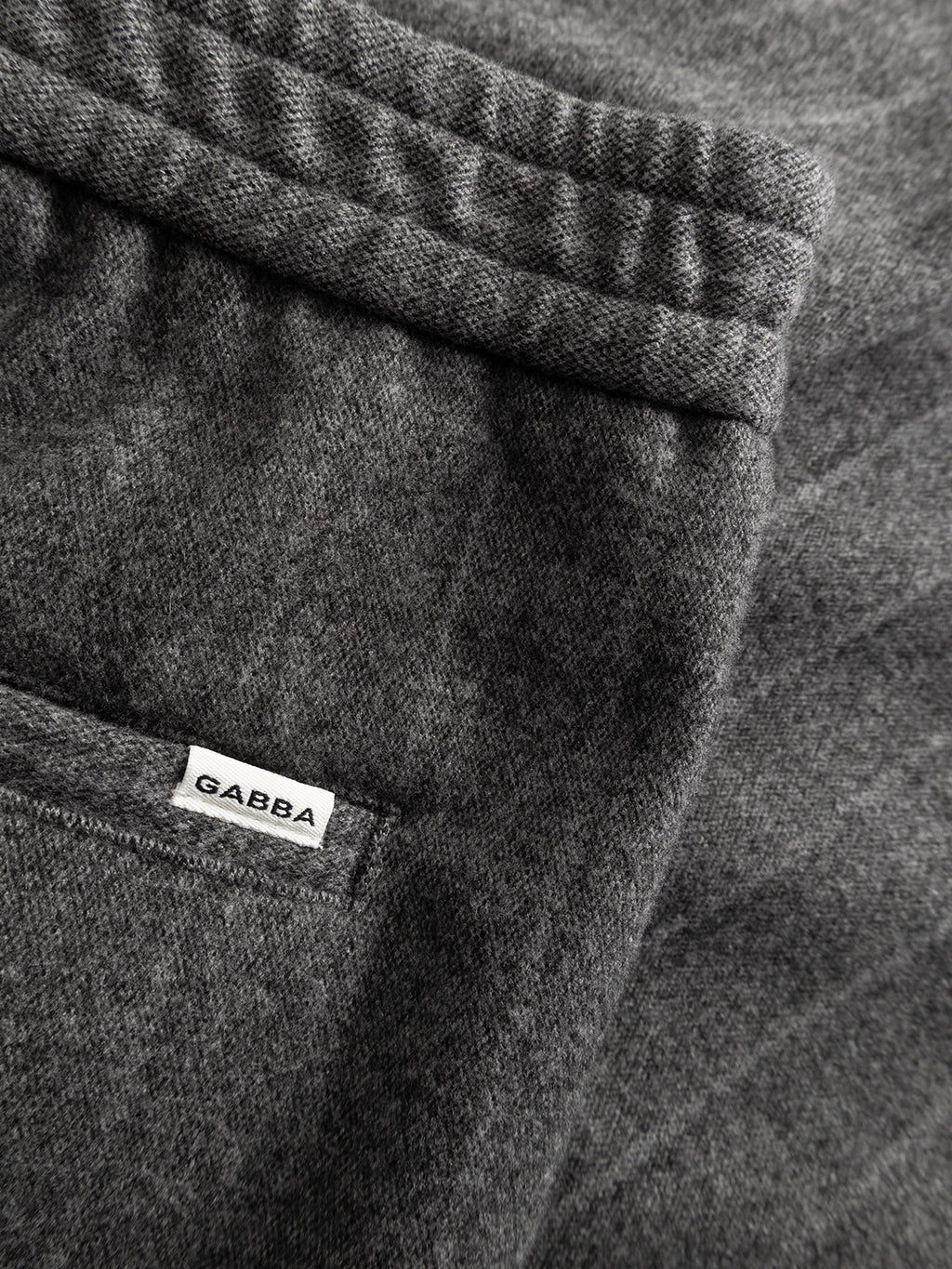 Close-up of dark grey Napoli 19 AX Stripes fabric, featuring visible stitching and a small white Gabba tag attached near a seam or pocket—showcasing the refined chino pocket construction and subtle details characteristic of relaxed fit pants by Gabba.