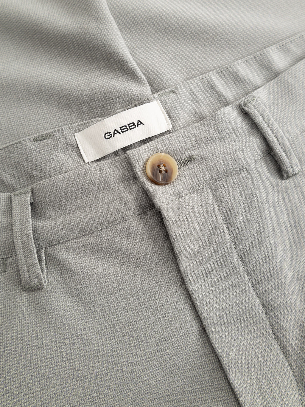 Close-up of the GABpaul Deme Pant by Gabba in Mineral Gray—a slim-fit style with a tan button, belt loops, and a white inner waistband label featuring the Gabba brand name.
