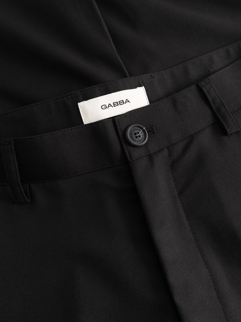 Close-up of GABjoe Turnup Fin Pant - Black by Gabba: classic tapered-fit black trousers with visible waistband, black button, belt loops, and inner white Gabba label. Made from a durable and comfortable twill viscose blend.