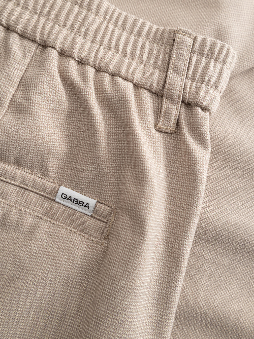 Close-up of the GABmonza Tasca Deme Pant in Birch by Gabba—a light beige linen pant with a relaxed straight fit, elastic waistband, belt loops, back pocket, and a white Gabba label above the pocket.
