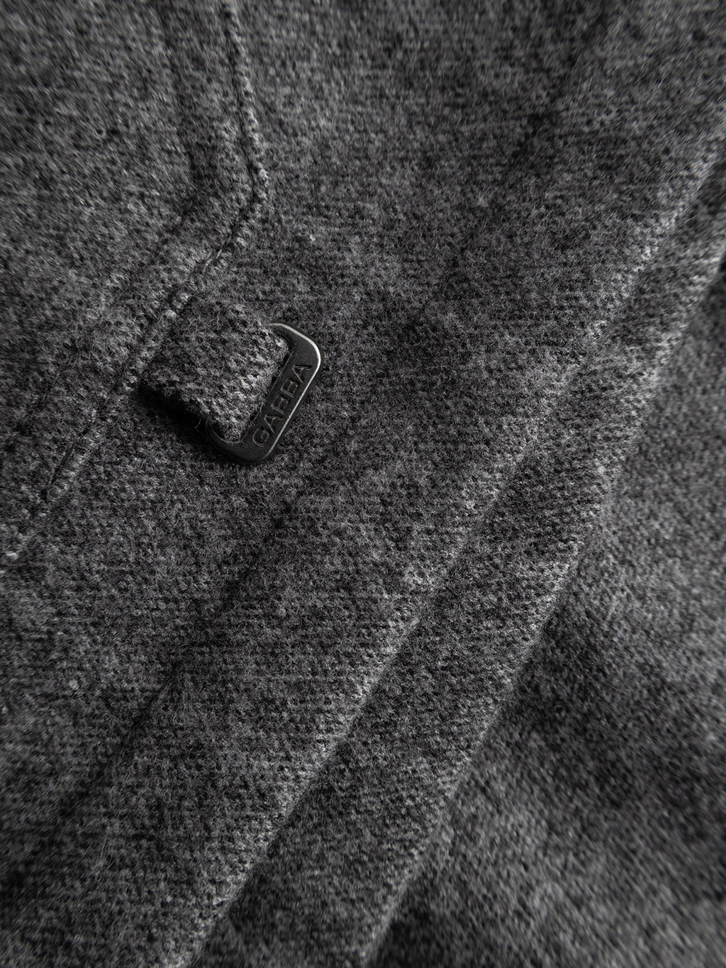 Close-up of a dark grey wool and viscose-blend fabric from the Gabba GABlibre AX Stripes LS Overshirt, featuring a metal tag with the brand name near a stitched edge, suggesting part of this boxy fit garment.