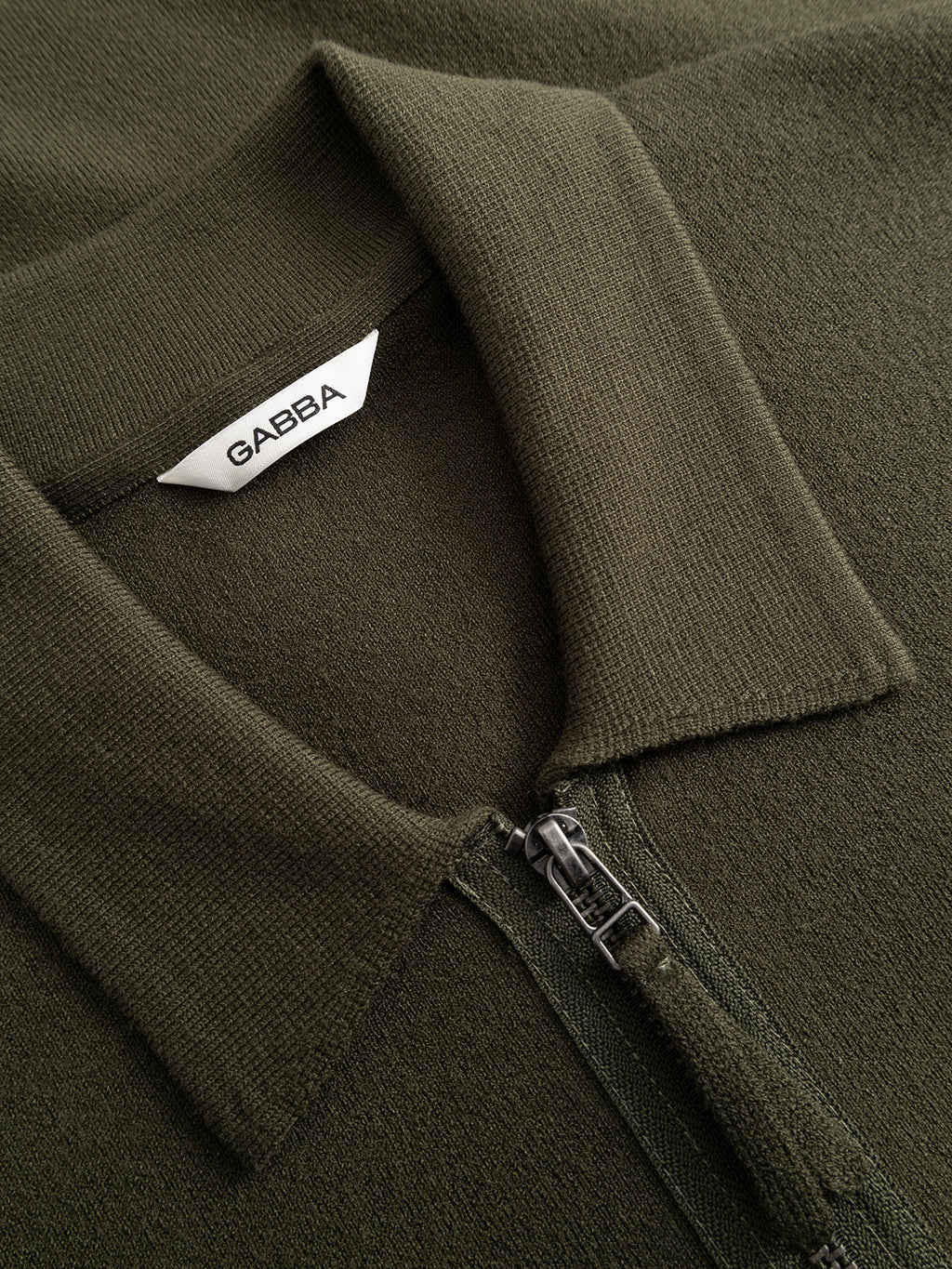 Close-up of the Gabba GABadriel Zip Lume LS Knit in Forest Night, a regular-fit olive green zip-up collared shirt with a visible Gabba label inside the collar. The viscose blend fabric’s texture and full zip closure details are clearly highlighted.