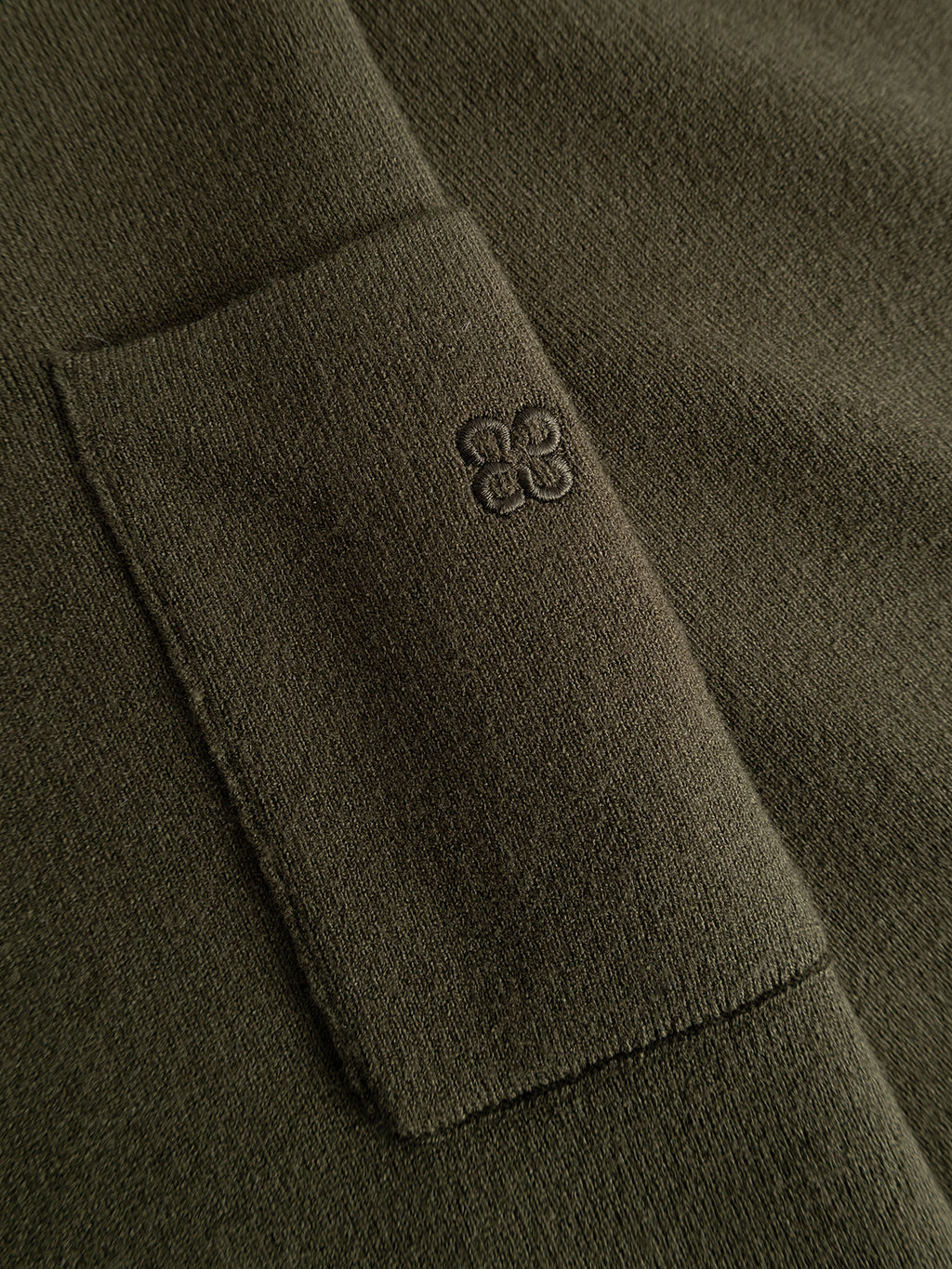Close-up of the Gabba GABadriel Zip Lume LS Knit in Forest Night, showing its regular-fit olive green viscose-blend knit fabric with a soft texture. Features include a small embroidered four-leaf clover near the square pocket seam and visible stitching details.
