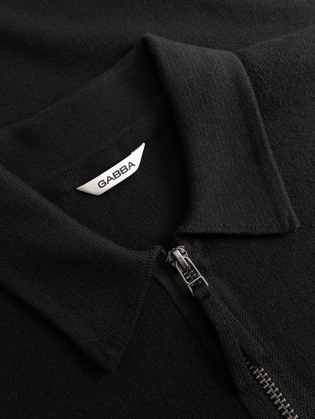 Close-up of the GABadriel Half Zip Lume LS Knit in Meteorite by Gabba, a black, regular-fit knit collared shirt featuring a partially open zipper that reveals a white label inside the collar reading Gabba.