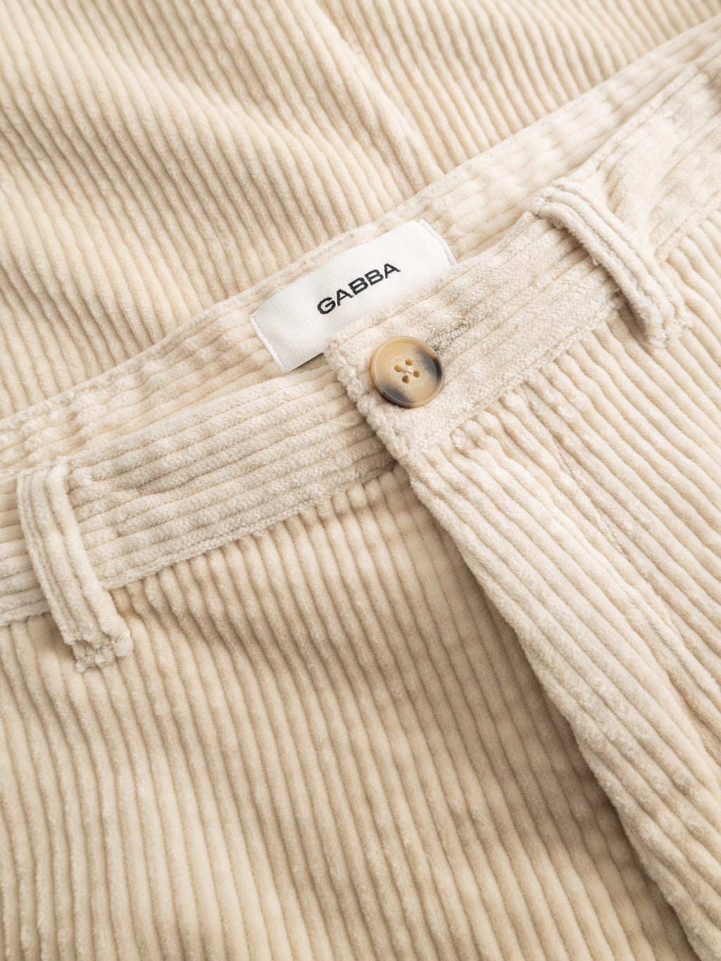 Close-up of the GABjoe Nopleats Ibre Pant in Birch by Gabba, featuring a beige tapered fit in stretch corduroy. Details include a button, belt loop, and a white label with Gabba text. The soft, ribbed fabric highlights characteristic corduroy texture and displays neat stitching and a clean finish.
