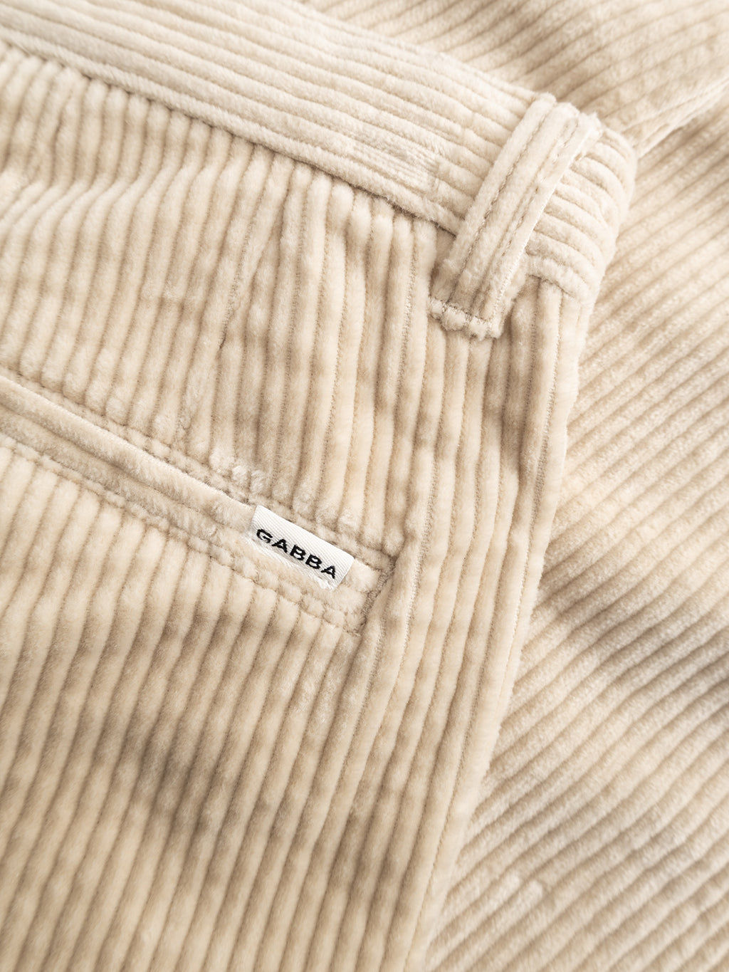 Close-up of the GABjoe Nopleats Ibre Pant in Birch by Gabba, showcasing the beige stretch corduroy fabric with its distinct textured ridges. The classic chino construction is highlighted by a visible pocket and a small white Gabba label, emphasizing the pant’s timeless yet comfortable style.