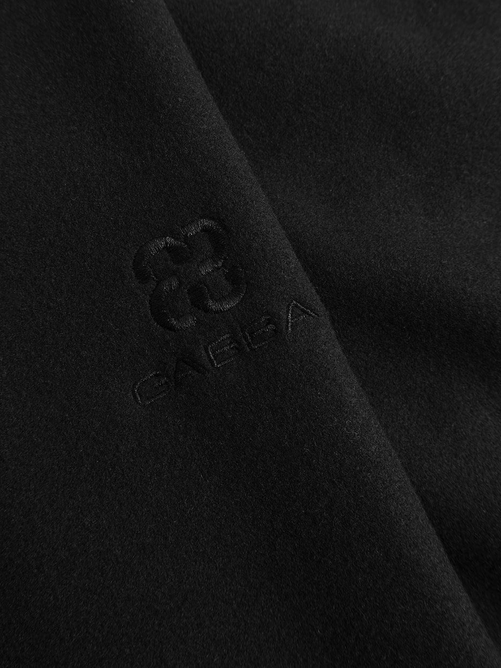 Close-up of black, wool-inspired fabric featuring a subtle, embroidered floral-like logo above the word Gabba—all stitched in matching black thread—creates a monochromatic, textured design on the GABtheo City Jacket in Black. This versatile piece from Gabba is perfect for everyday layering or styling with your favorite overshirt jacket.