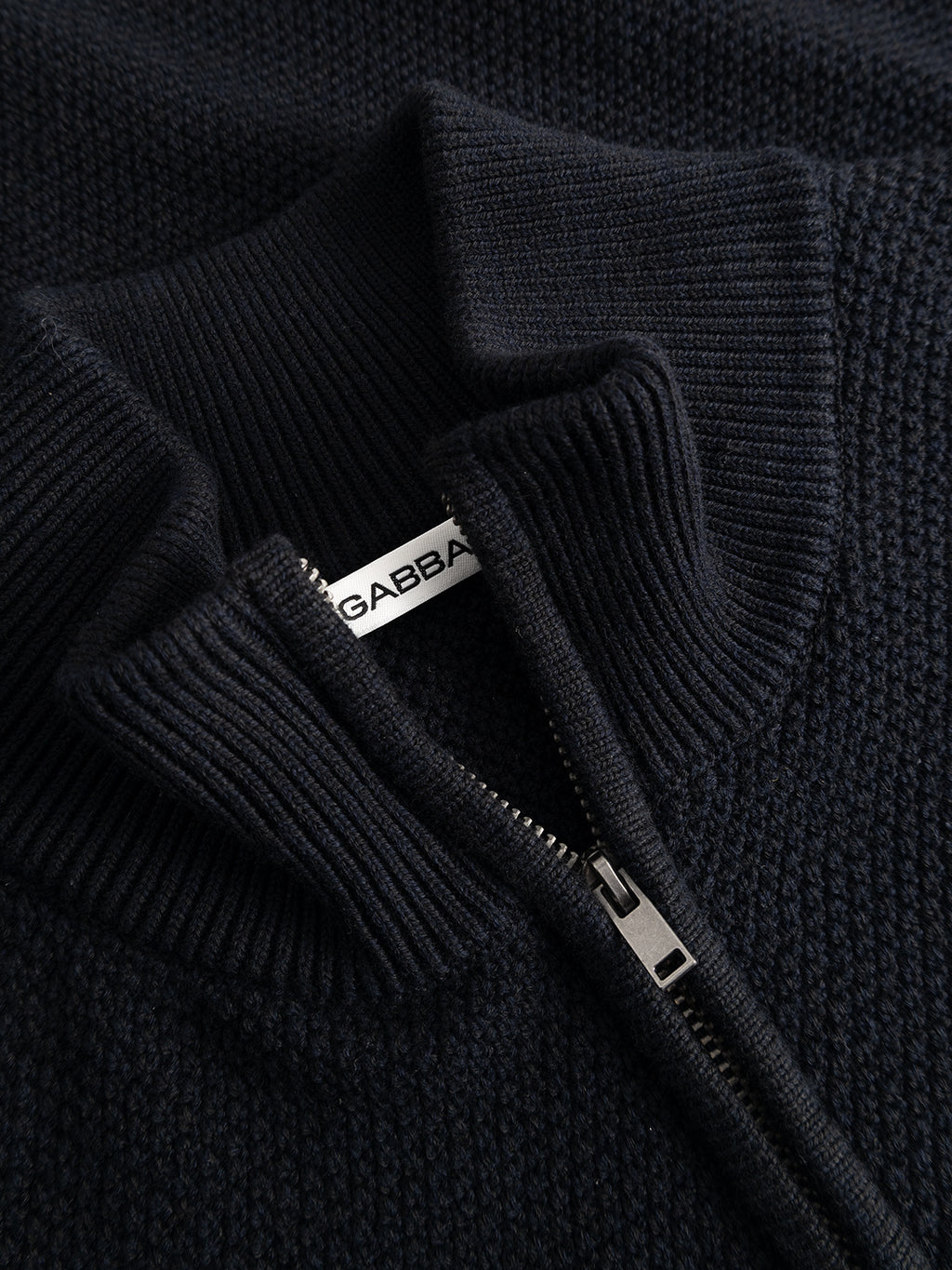 Close-up of the GABlamix Coro Half LS Knit in Blue Black by Gabba, featuring a textured, dark cotton-blend knit and a partially open silver zipper that reveals a white Gabba tag. The ribbed, folded half-zip collar highlights the garment’s quality craftsmanship and regular fit silhouette.