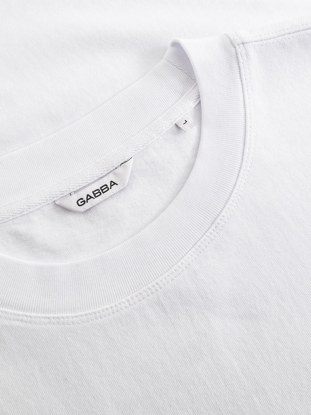 Close-up of the GABlegin Wix Boxy Hour SS T-shirt in white by Gabba, crafted from soft cotton jersey. The image highlights the collar, neckline stitching, and a white fabric tag with “Gabba” in black text. Featuring a relaxed boxy fit and a subtle embroidered Gabba logo, this everyday essential offers contemporary style.