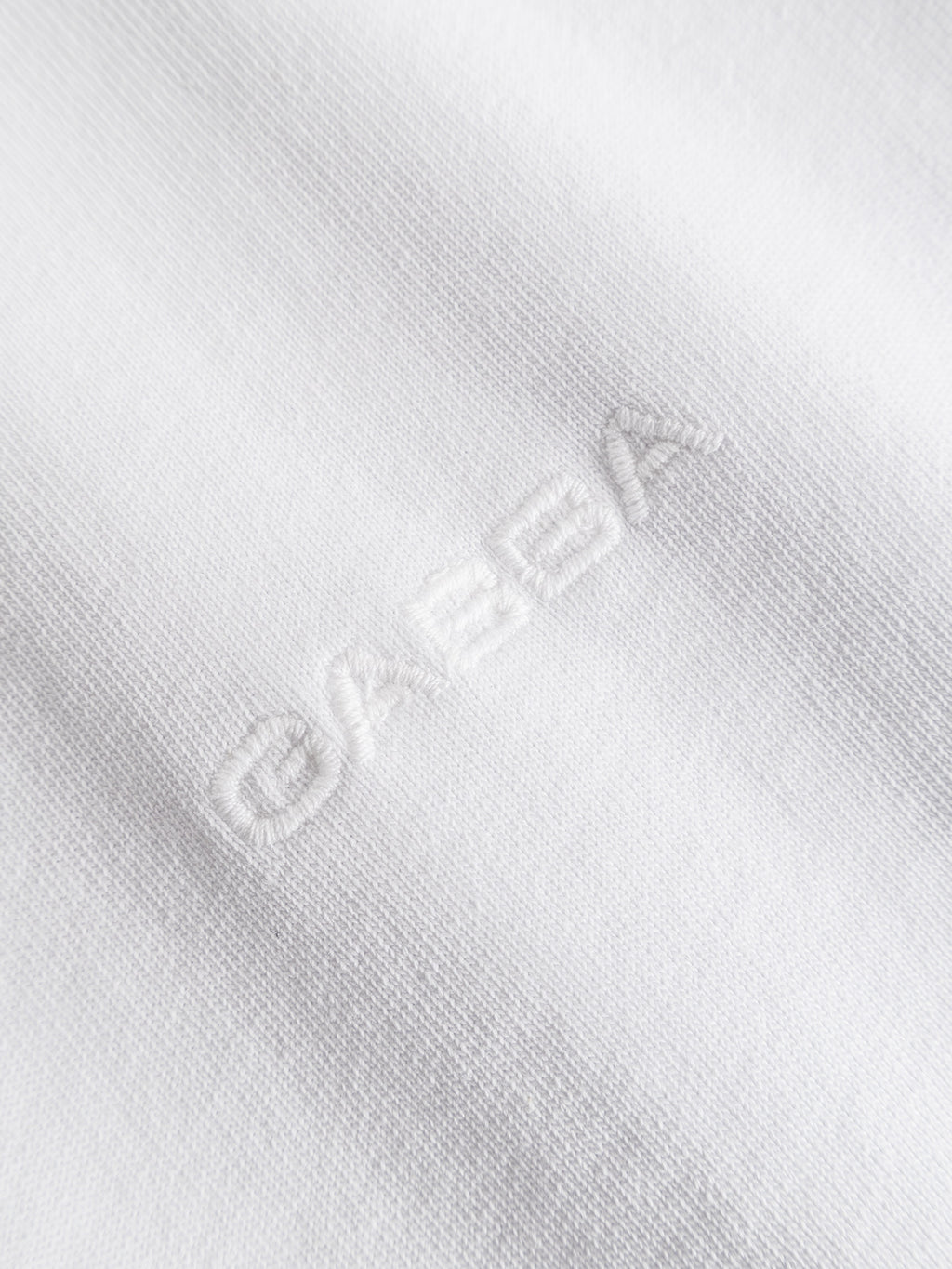 Close-up of the white cotton jersey fabric of the GABlegin Wix Boxy Hour SS T-shirt by Gabba, featuring “OREBA” subtly stitched in matching white thread for a textured text effect on the soft material. The relaxed, boxy fit is finished with an understated embroidered Gabba logo, adding a refined touch.