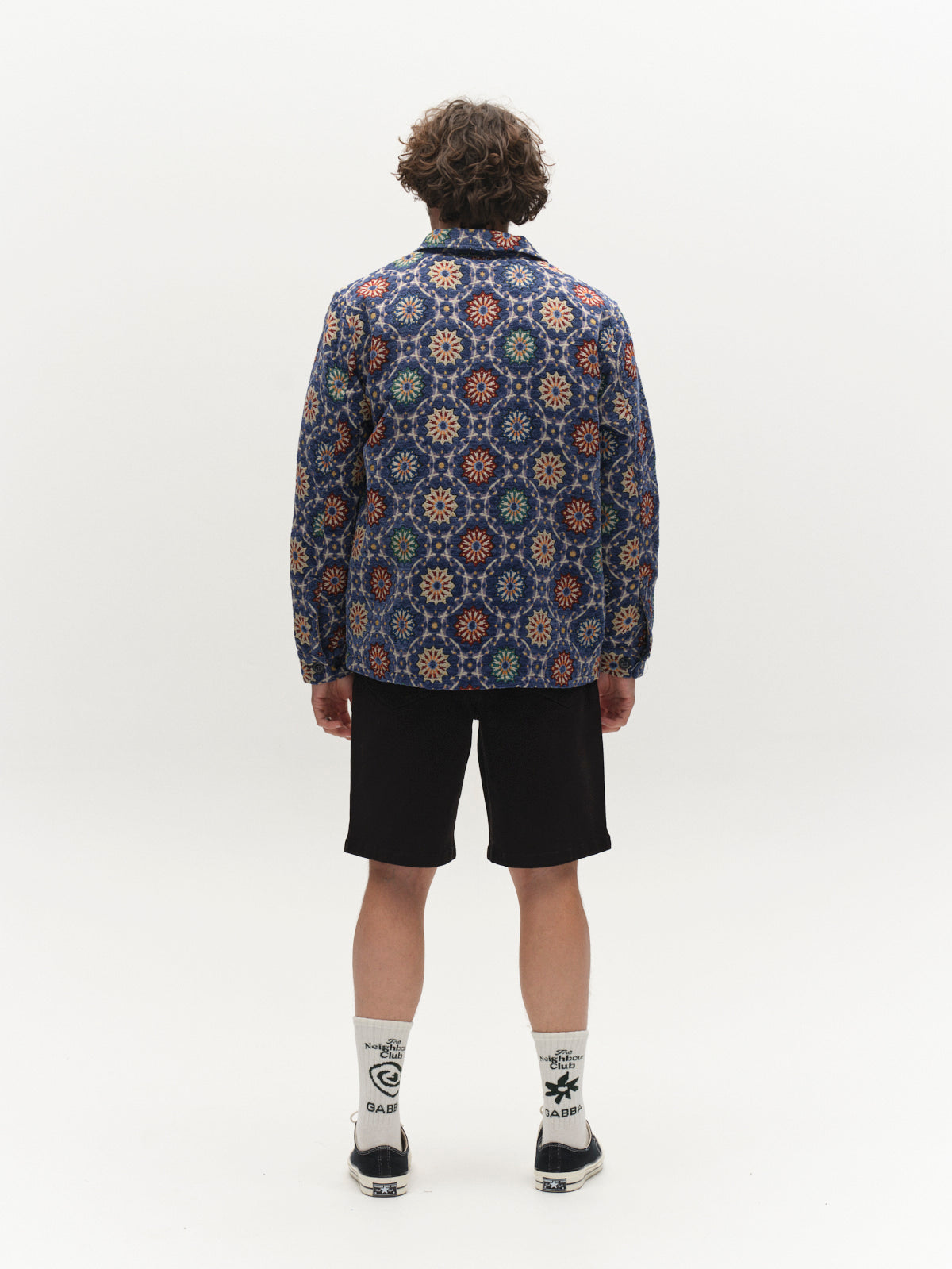 A person with curly hair stands facing away, wearing the Gabba GABsimone Nava LS - Multi Pattern shirt with a resort collar, paired with black shorts, white graphic socks, and black shoes against a plain white background.
