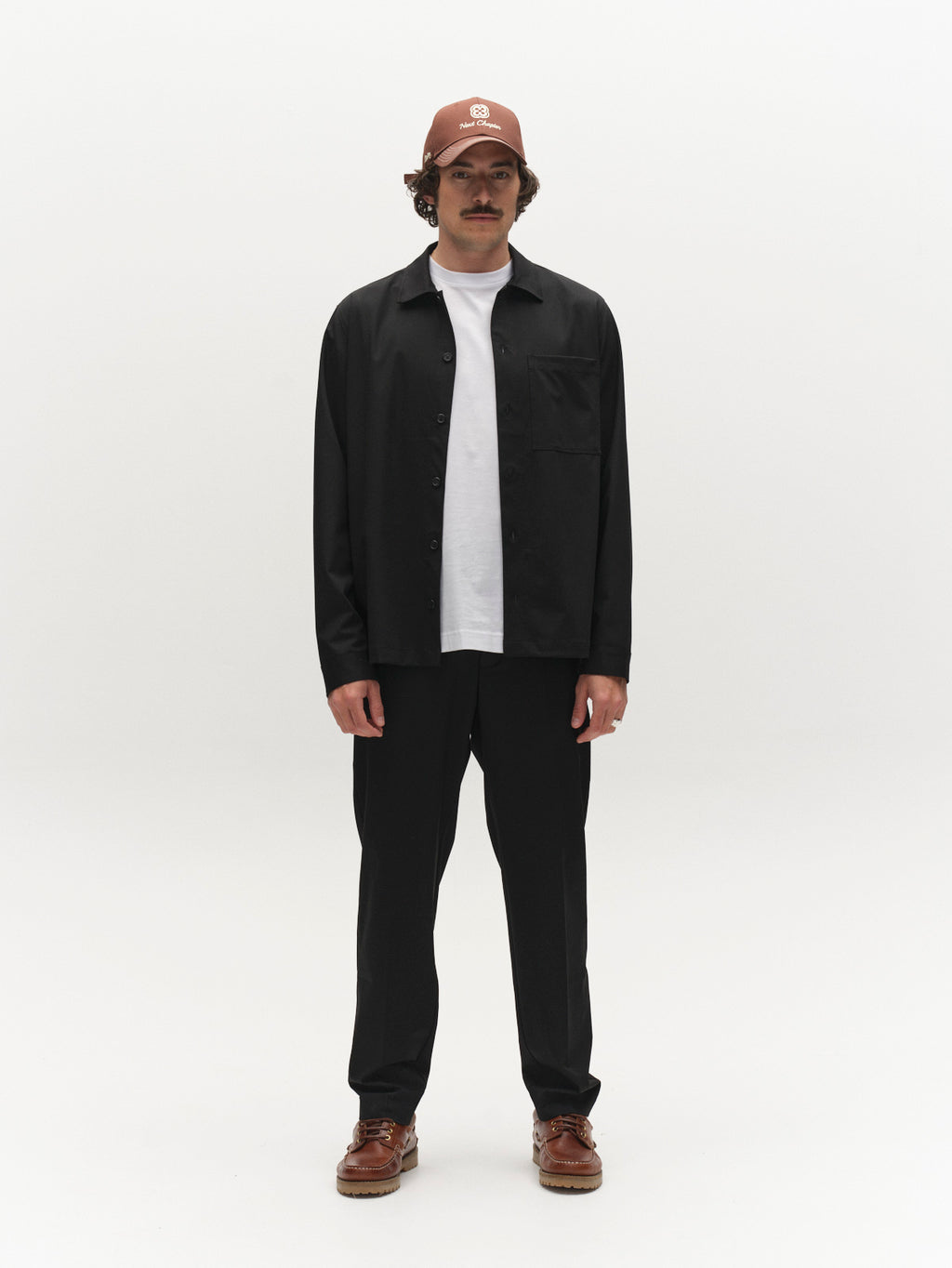 A man stands facing forward against a plain white background, wearing a brown cap, the Gabba GABtogger Tailor Dressy Overshirt in black over a white t-shirt, regular-fit black pants, and brown shoes.