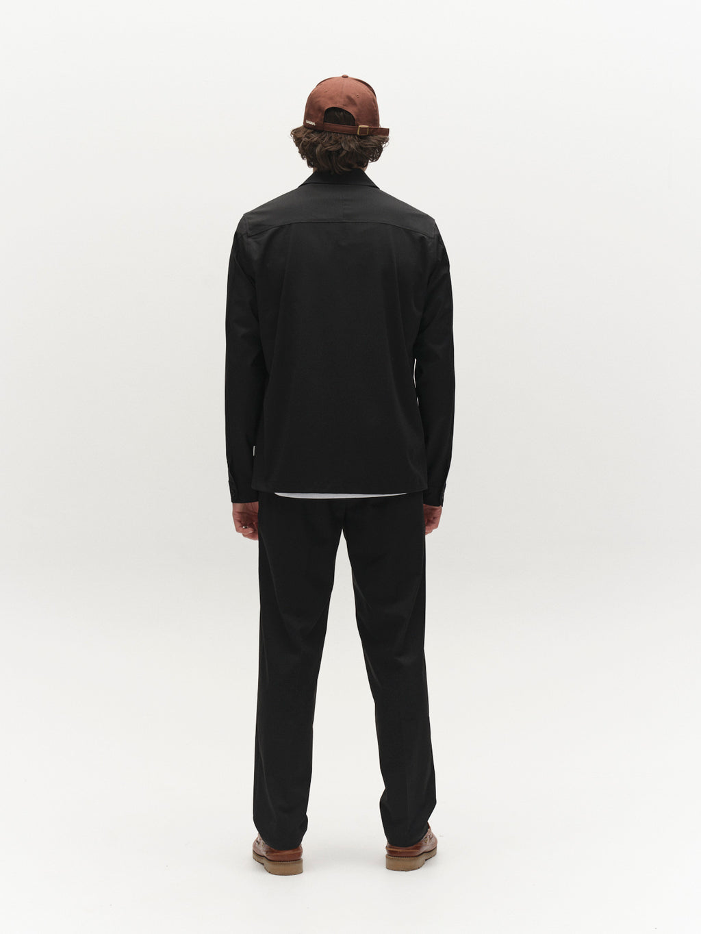 A person stands facing away against a plain white background, wearing a brown cap, black pants, tan shoes, and the Gabba GABtogger Tailor Dressy Overshirt in black.