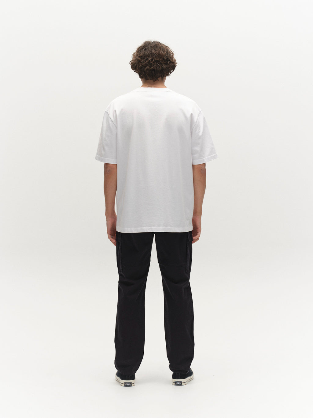 A person with short, curly hair stands facing away, wearing the Gabba GABnigel Wix Pkt Boxy Hour SS T-Shirt in white—a heavyweight cotton tee featuring a subtle chest pocket—paired with black pants and sneakers against a white background.