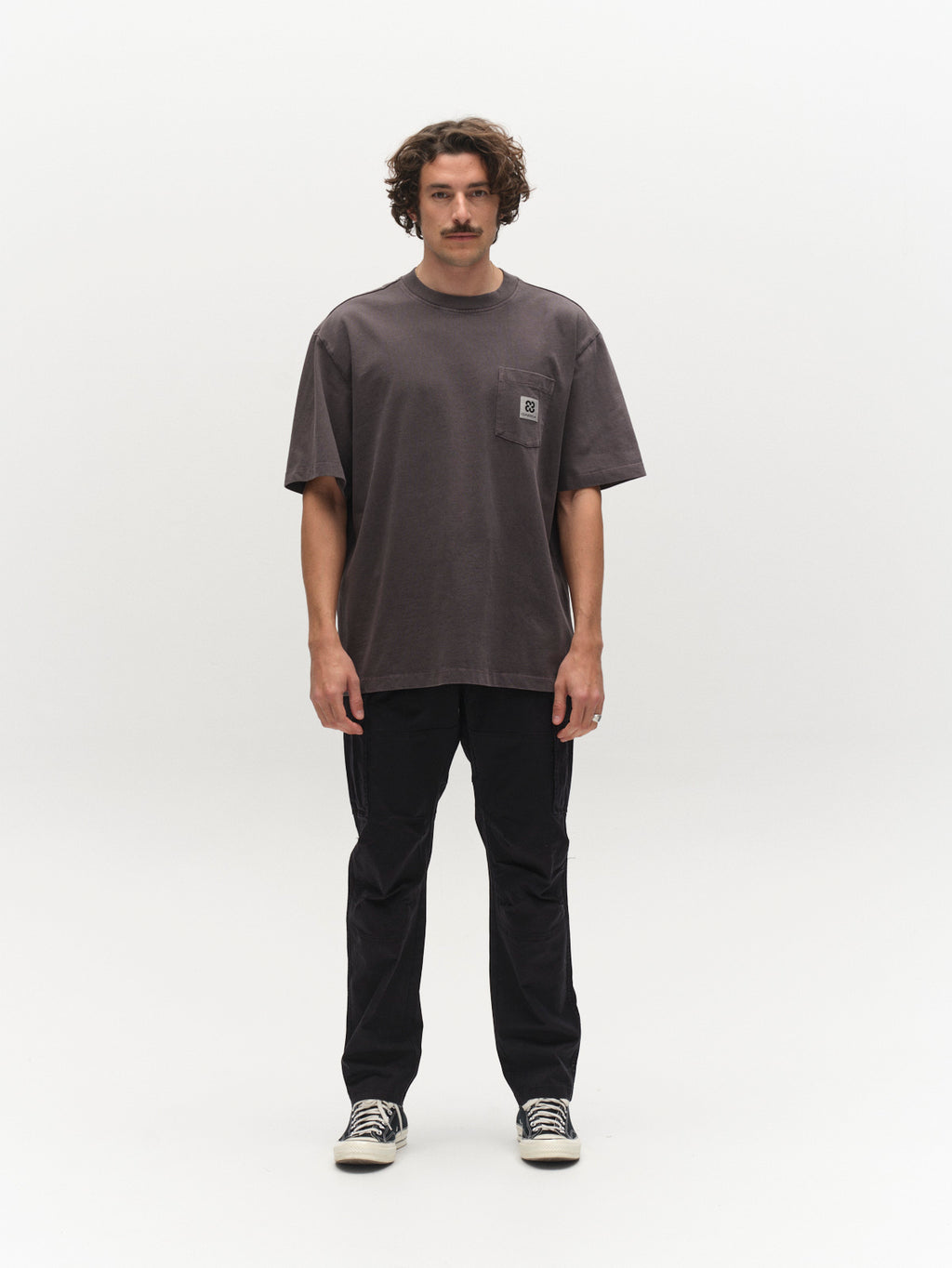 A man with curly hair and a mustache stands facing forward against a plain white background, wearing the Gabba GABnigel Wix Pkt Boxy Hour SS T-Shirt in dark grey—a loose, boxy-fit tee crafted from heavyweight cotton with a small chest logo—paired with black pants and black sneakers.
