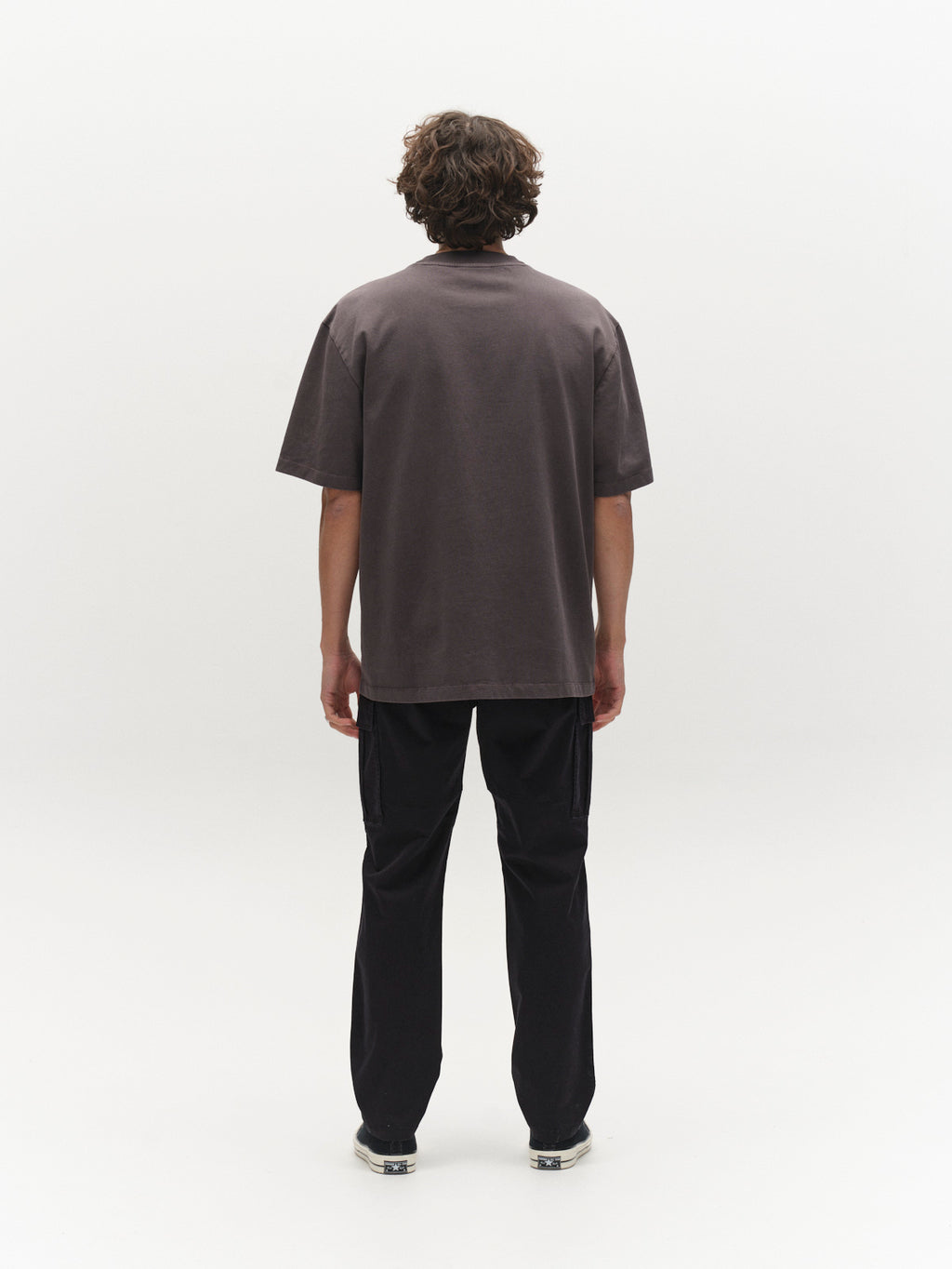 A person with curly hair stands facing away, wearing the Gabba GABnigel Wix Pkt Boxy Hour SS T-Shirt in dark grey—a loose, heavyweight cotton boxy tee with a chest pocket—paired with black pants and sneakers against a plain white background.