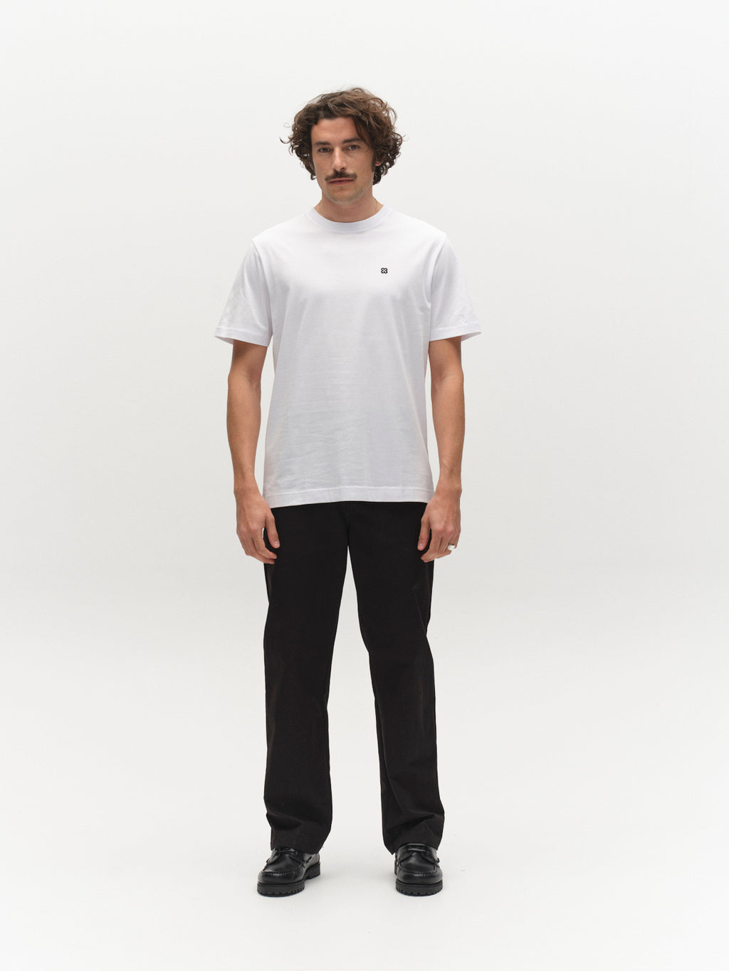 A man with curly hair and a mustache faces forward, wearing the Gabba GABdune Element GOTS SS T-Shirt in white, paired with black pants and shoes, standing against a white background.
