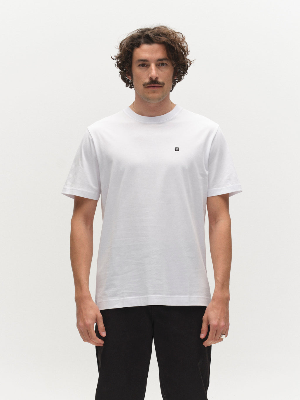 A man with curly hair and a mustache wears the Gabba GABdune Element GOTS SS T-Shirt in white, paired with black pants, standing against a white background.