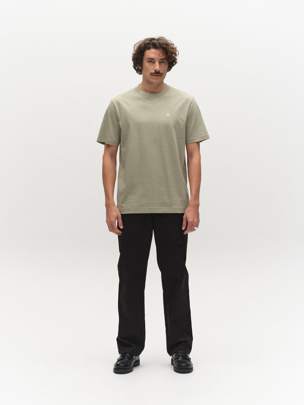 A man with wavy hair and a mustache stands facing forward against a plain white background, wearing the Gabba GABdune Element GOTS SS T-Shirt in Light Green, paired with black pants and black shoes.