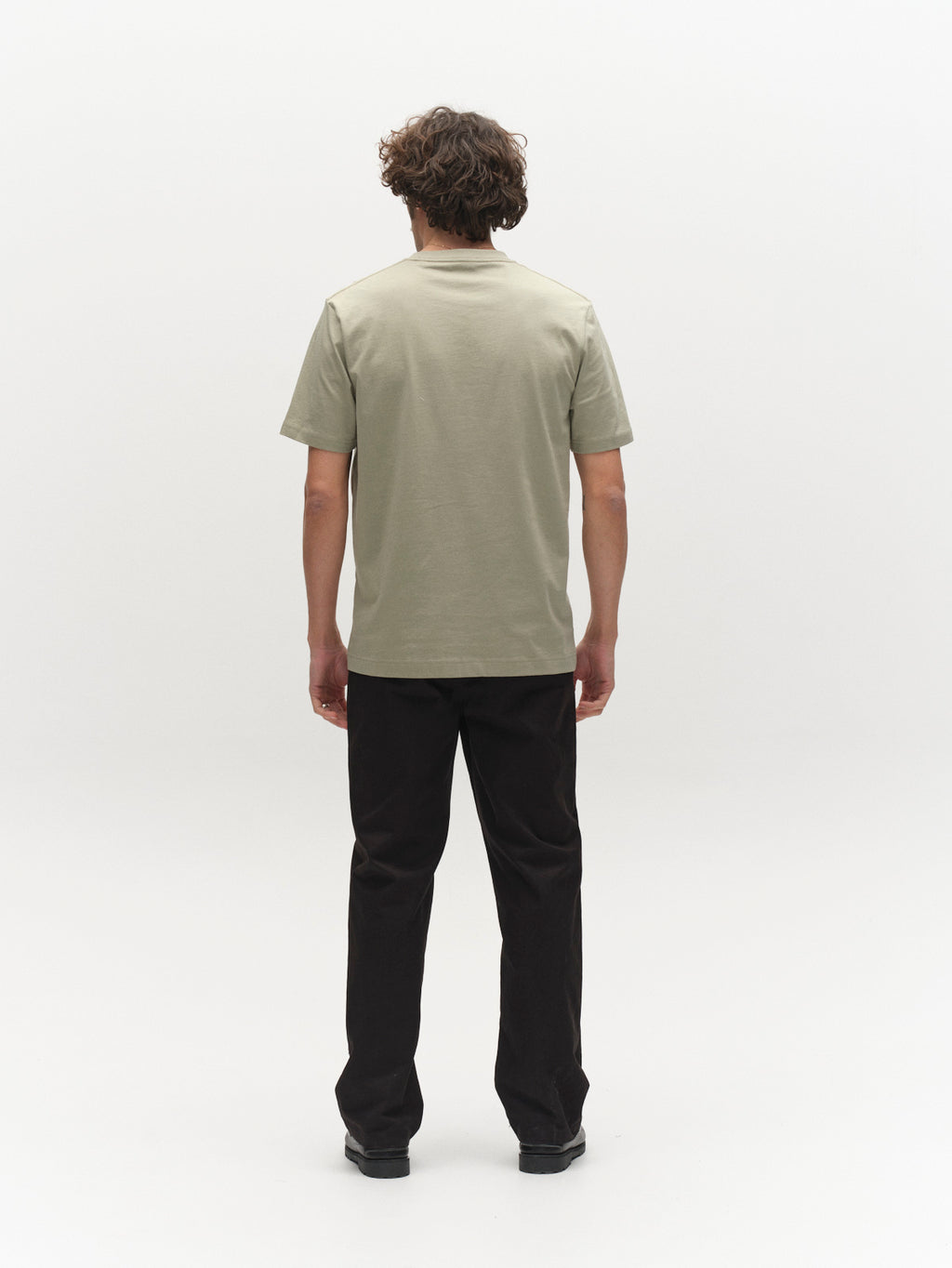 A person with curly hair faces away from the camera, wearing the Gabba GABdune Element GOTS SS T-Shirt in light green, paired with black pants and shoes, set against a plain white background.