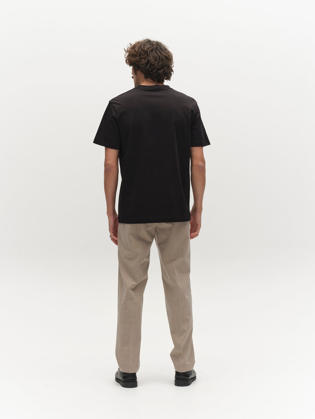 A person with curly hair stands facing away from the camera, wearing a Gabba GABdune Element GOTS SS T-Shirt in black, paired with beige pants and black shoes against a plain white background.