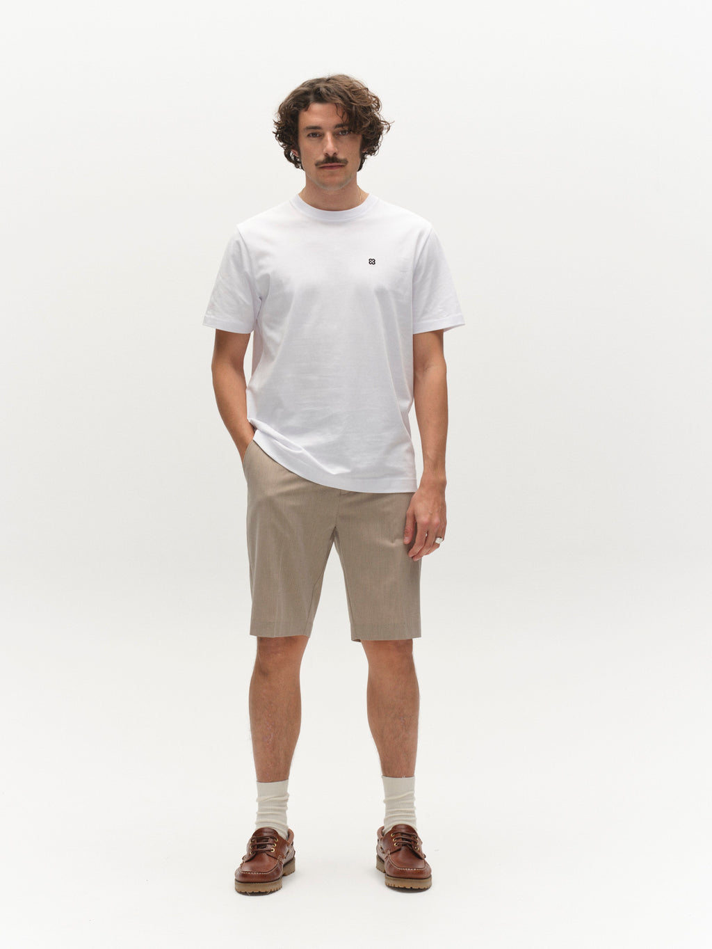 A man with wavy hair and a mustache stands against a white background wearing a white t-shirt, Gabba GABjoli Tailor Dressy Shorts in sand, white socks, and brown loafers. One hand is in his pocket as he faces the camera with a neutral expression.