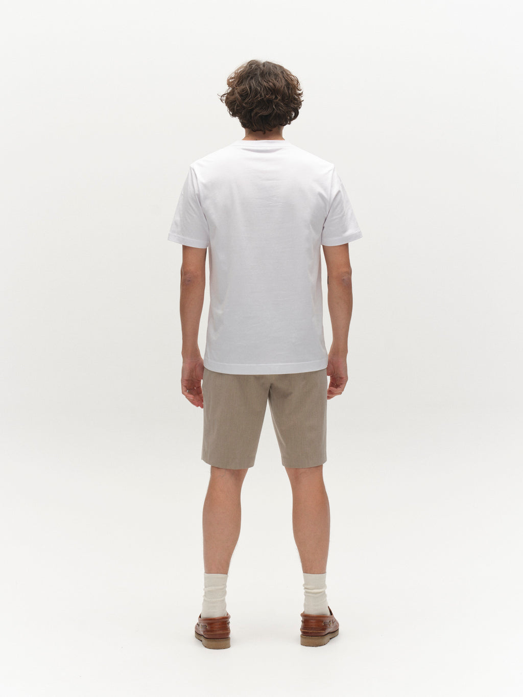 A person with short, curly brown hair stands facing away from the camera, wearing a plain white t-shirt, Gabba GABjoli Tailor Dressy Shorts in Sand, white socks, and brown shoes against a white background.
