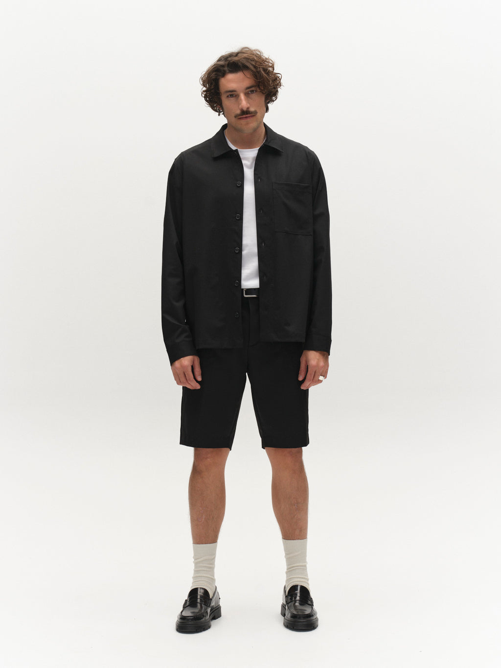 A man with curly hair and a mustache stands against a plain white background, wearing a black button-up shirt, Gabba GABjoli Tailor Dressy Shorts in black, white socks, black loafers, and a white t-shirt underneath.