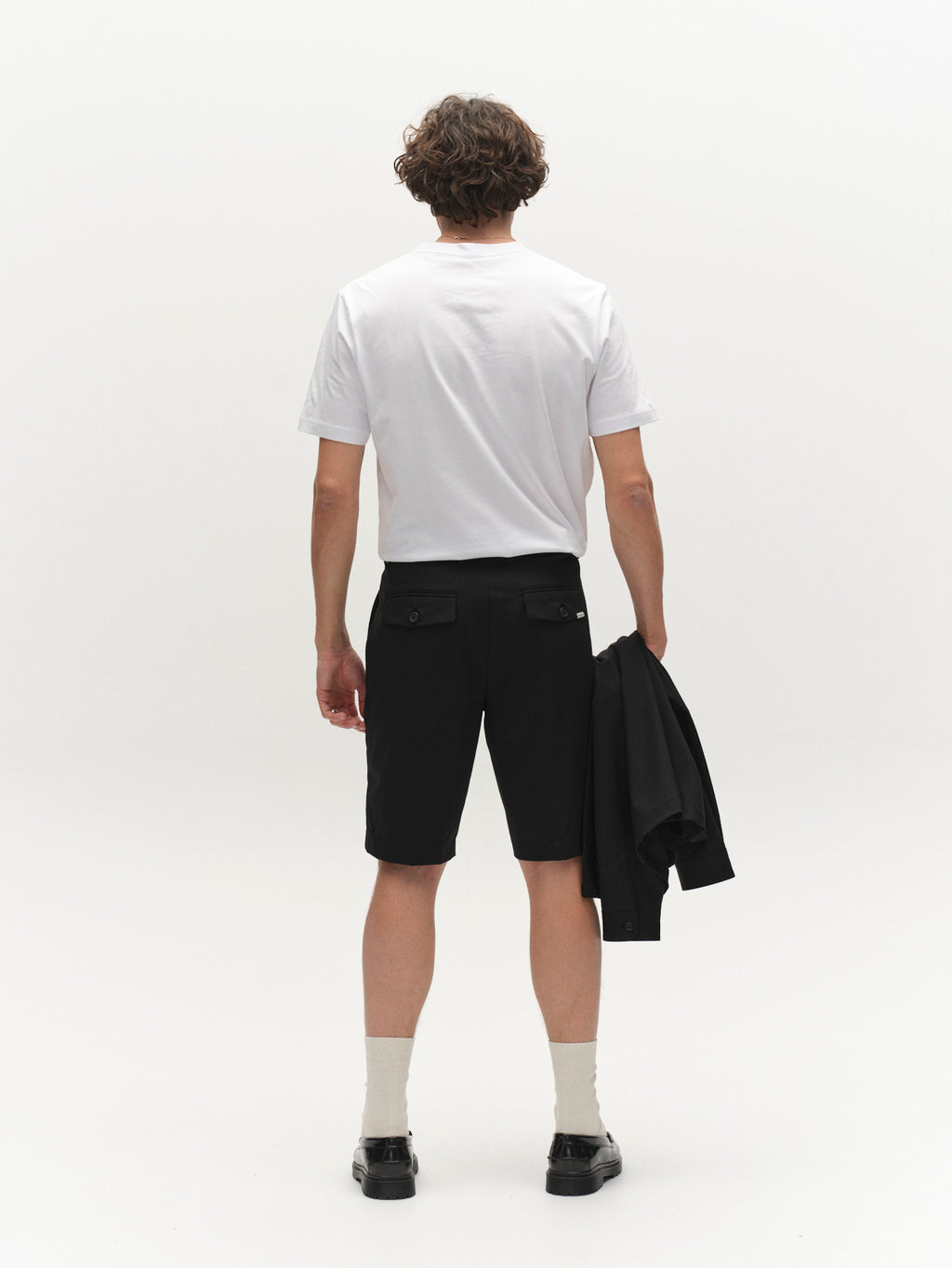 A person with short curly hair, in a white t-shirt, Gabba GABjoli Tailor Dressy Shorts - Black (relaxed fit), light socks, black shoes, faces away holding a black jacket in their left hand against a plain white background.