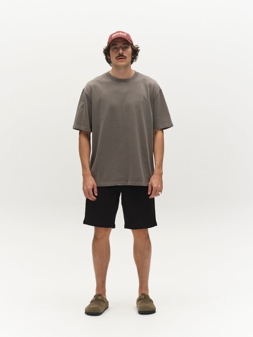 A man with a mustache stands facing forward against a white background, wearing a Gabba GABnigel Wix Boxy Hour SS t-shirt in Castor Gray, a red cap, black shorts, and brown slip-on shoes. His arms are relaxed at his sides.