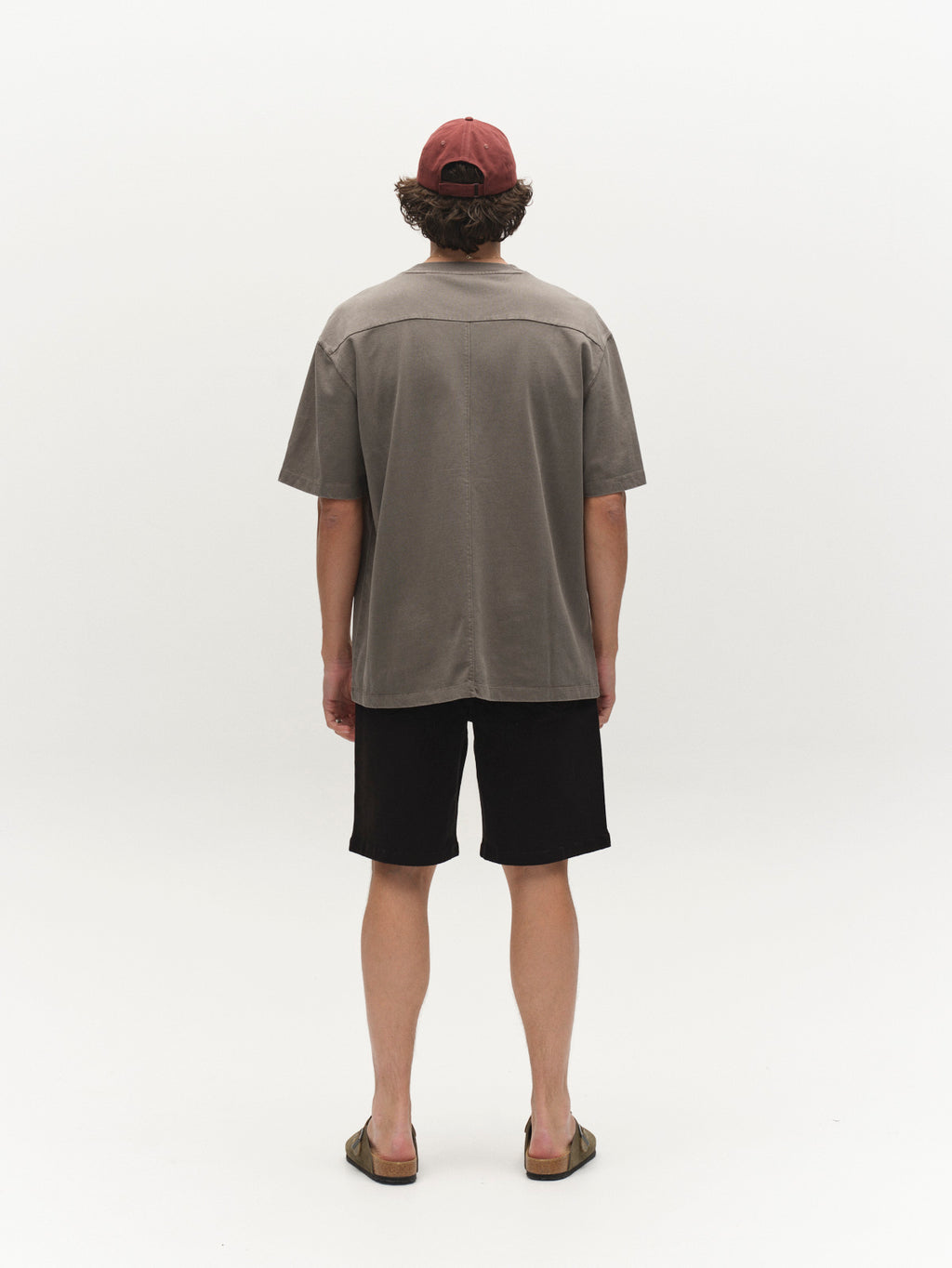 A person with curly hair, wearing a red cap, Gabba's GABnigel Wix Boxy Hour SS in Castor Gray, black shorts, and sandals stands facing away against a plain white background.