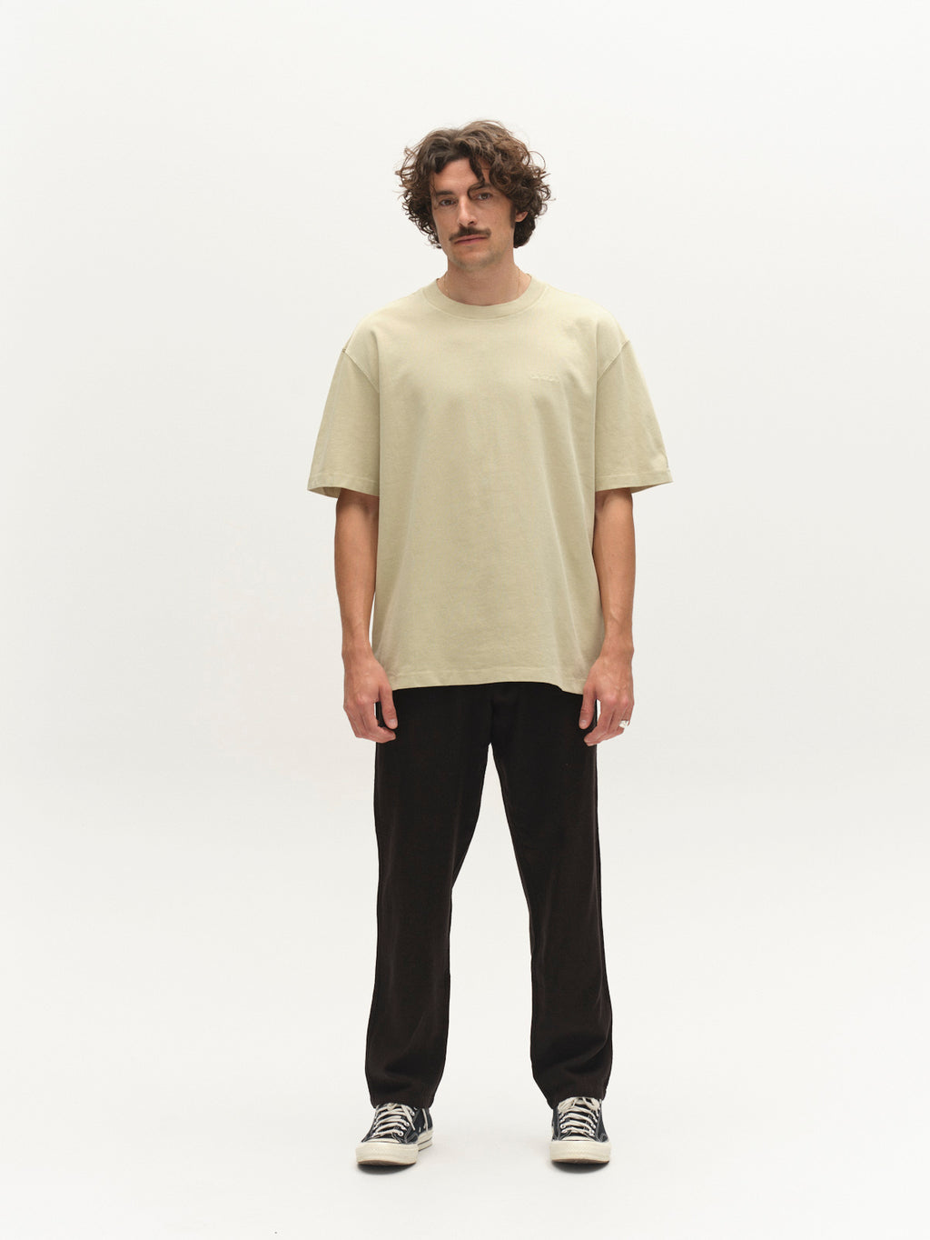 A man with curly brown hair and a mustache stands against a white background, facing forward in a neutral pose. He wears the Gabba GABnigel Wix Boxy Hour SS t-shirt in Alfa Green, black pants, and black-and-white sneakers.