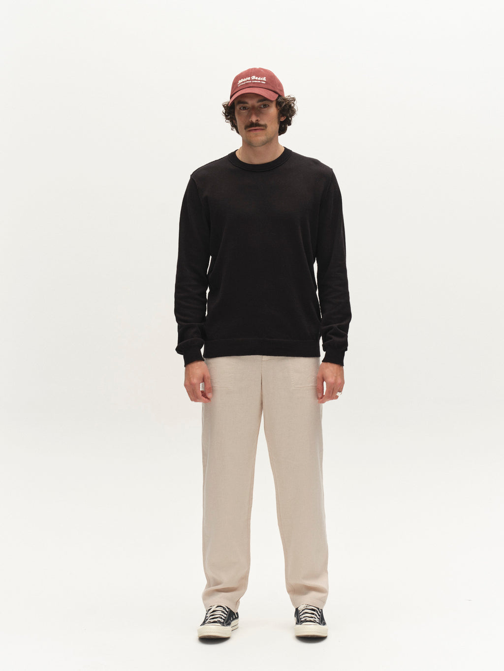 A man stands facing forward, wearing breathable Gabba GABzeta Lino - Birch pants, a black long-sleeve shirt, a red cap, and black-and-white sneakers against a plain white background.