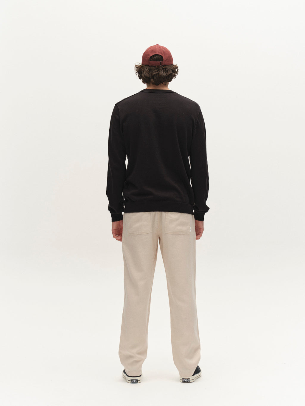 A person stands facing away from the camera, wearing a red cap, a black long-sleeve shirt, dark shoes, and Gabba GABzeta Lino - Birch pants against a plain white background.