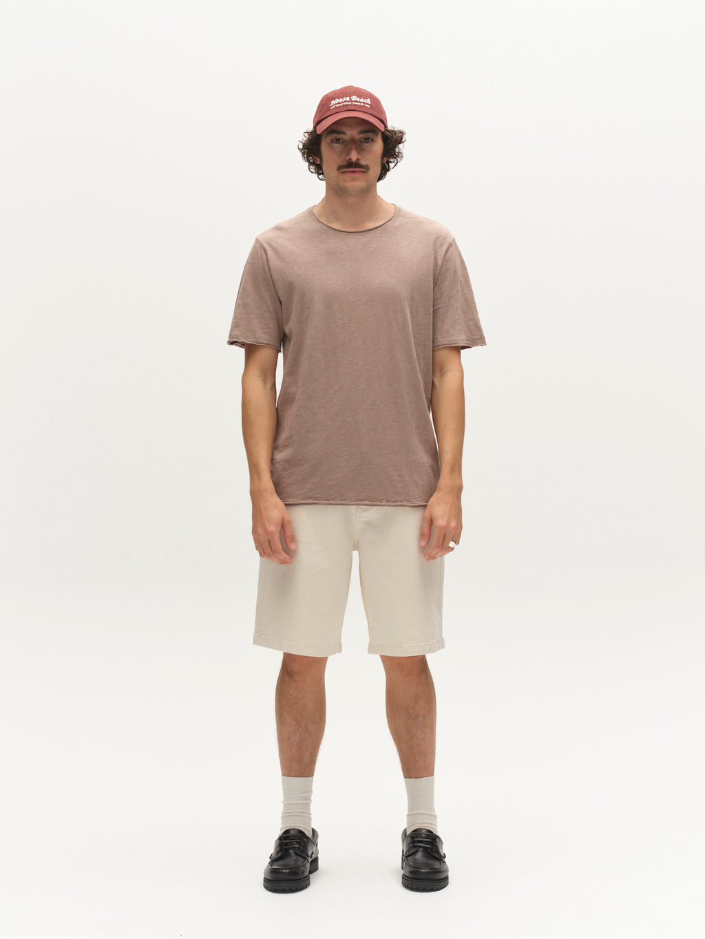 A man with a mustache stands facing forward in a maroon cap, beige shorts, socks, black shoes, and a Gabba GABkostant New SS - Fungi light brown 100% cotton slub fabric t-shirt against a plain white background.