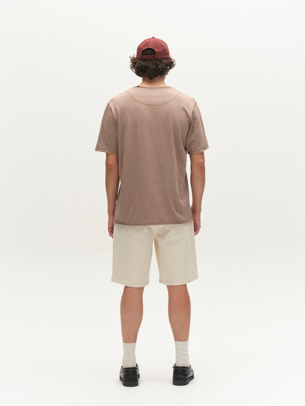 A person stands facing away from the camera on a plain white background, wearing the Gabba GABkostant New SS - Fungi brown slub fabric T-shirt with light beige shorts, white socks, black shoes, and a red cap.