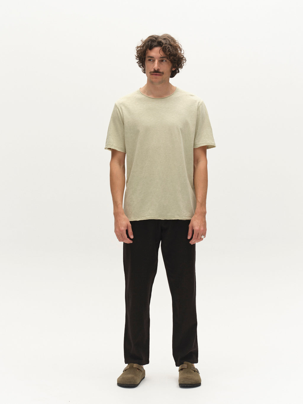 A man with curly brown hair and a mustache stands facing forward, wearing the Gabba GABkostant New SS - Alfa Green t-shirt, black pants, and brown shoes against a plain white background.