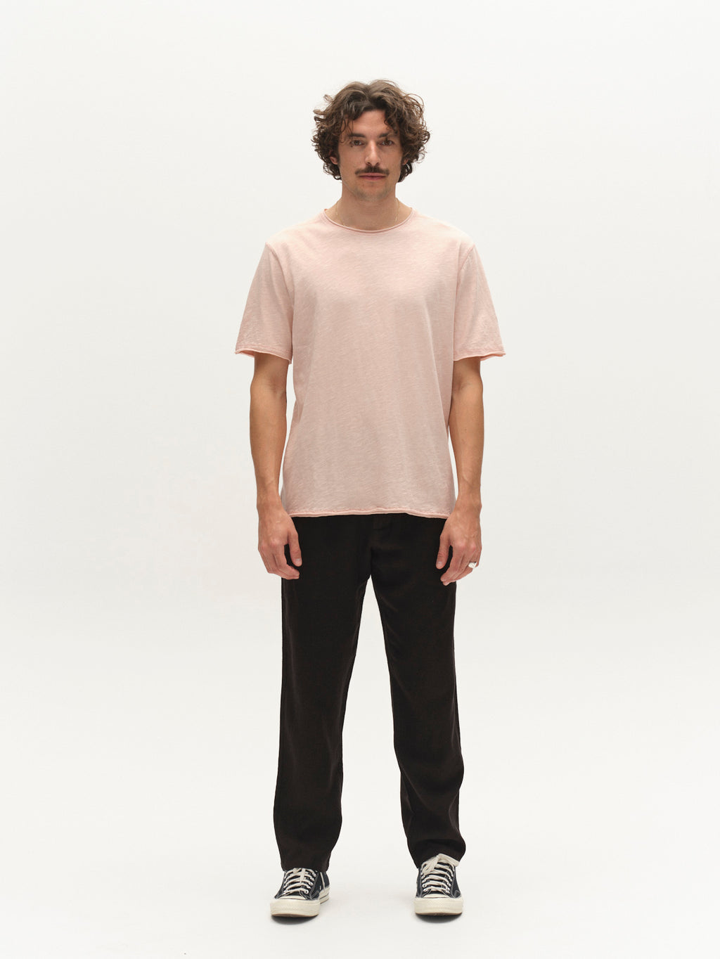 A man with curly hair and a mustache stands against a white background, wearing the Gabba GABkostant New SS t-shirt in Misty Rose, paired with black pants and black-and-white sneakers. His arms are relaxed at his sides.