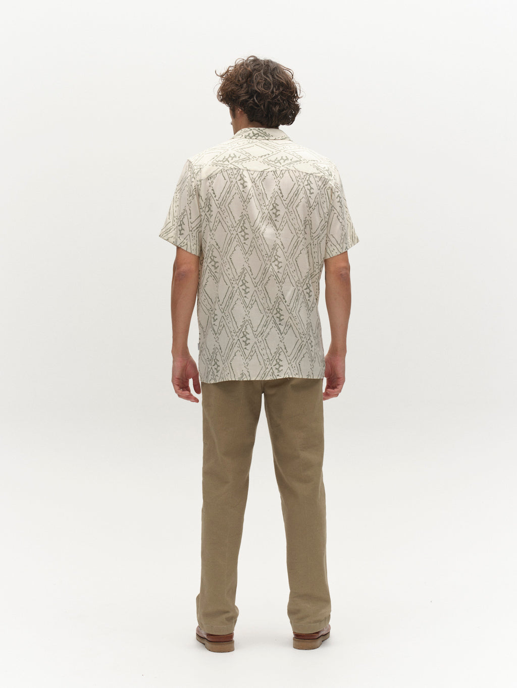 A person with curly hair faces away, wearing the Gabba GABsven Reg Zag SS Shirt in Multi Pattern, made from a TENCEL cotton blend, and tan pants against a plain white background.