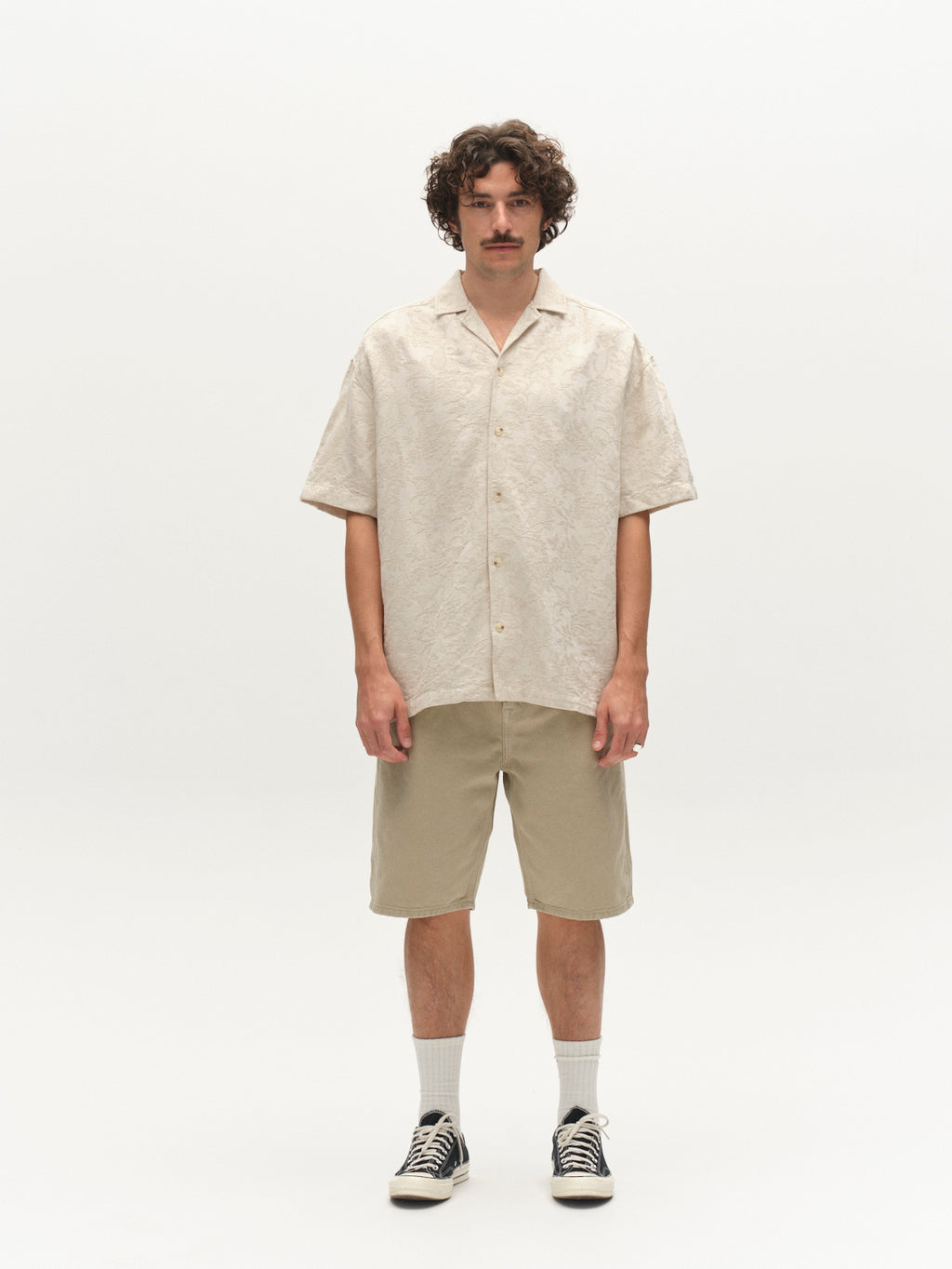 A man with curly hair and a mustache stands against a plain white background, wearing the GABjens Over Resort Soft SS Shirt in Ecru by Gabba, tan knee-length shorts, white socks, and black sneakers.