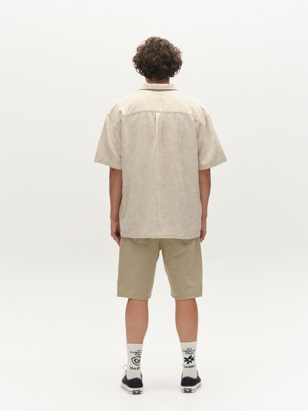A person with curly hair stands facing away, wearing the Gabba GABjens Over Resort Soft SS Shirt in Ecru, paired with khaki shorts, white graphic socks, and black sneakers against a plain white background.