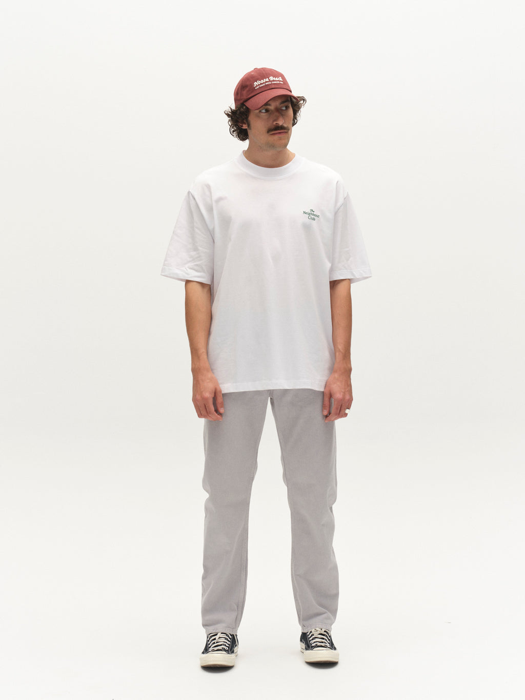 A person with a mustache, wearing a red cap, white oversized t-shirt, Gabba GABcarl Antiq Jeans in denim blue, and black-and-white sneakers, stands against a plain white background looking slightly to the side.