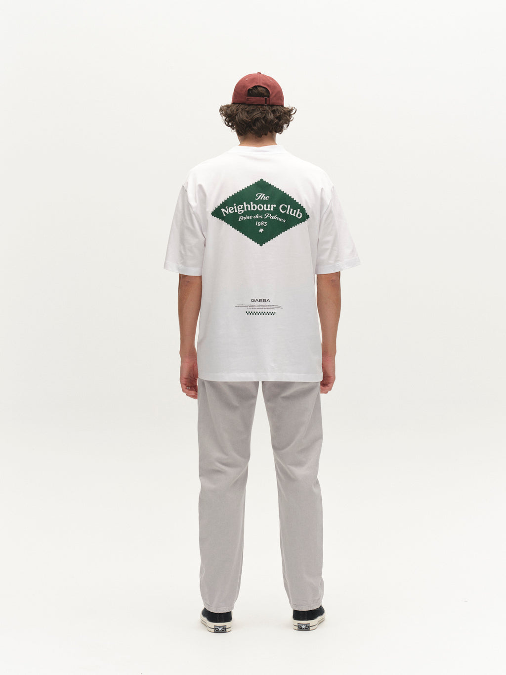 A person stands facing away, wearing a maroon cap, white graphic tee with green Neighbour Club logo on the back, Gabba GABcarl Antiq Jeans in denim blue, and black shoes against a plain white background.