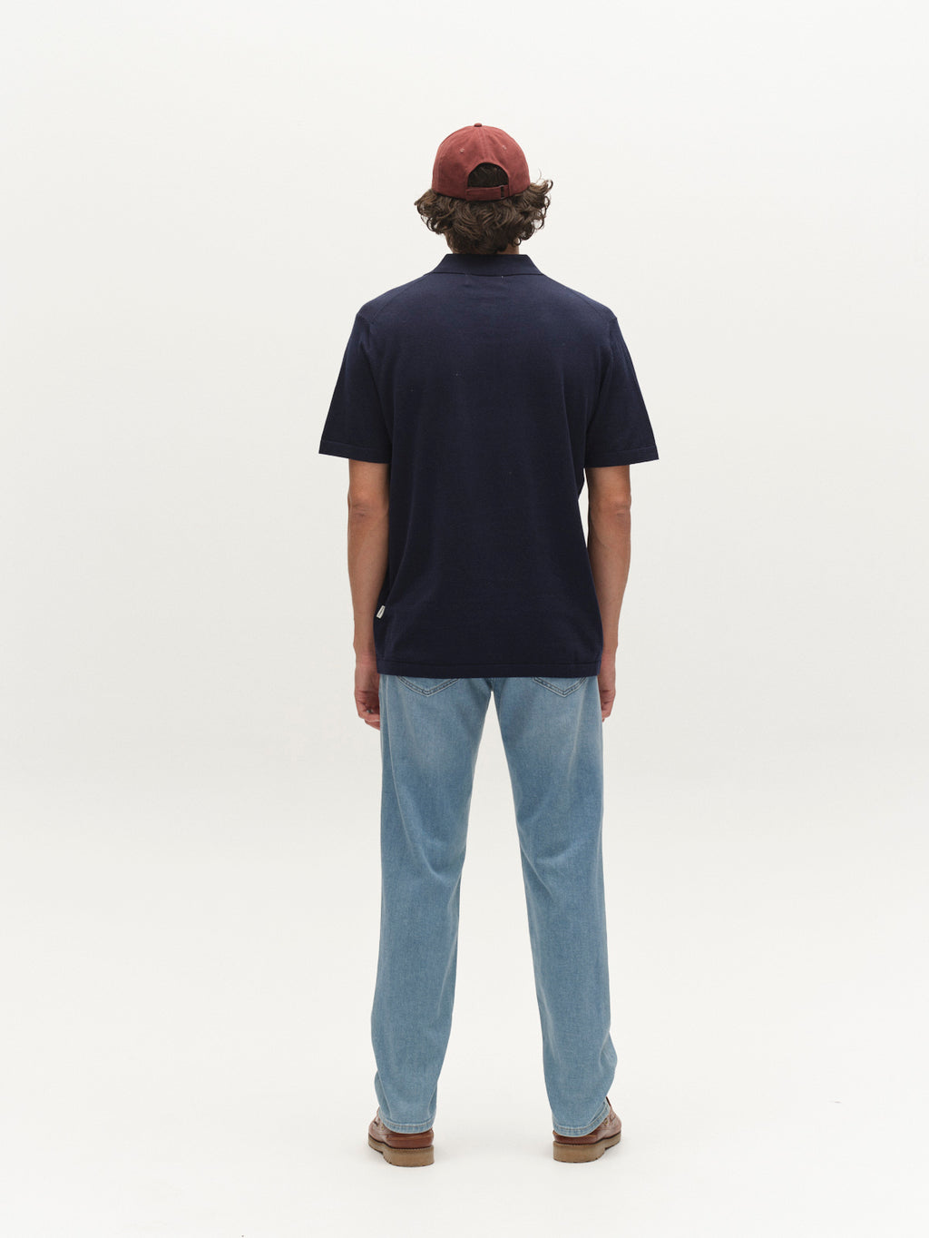 A person stands facing away in a maroon cap, navy polo, Gabba GABmarc Hobok Jeans in mid blue denim made with regenerative cotton, and brown shoes against a plain white background.